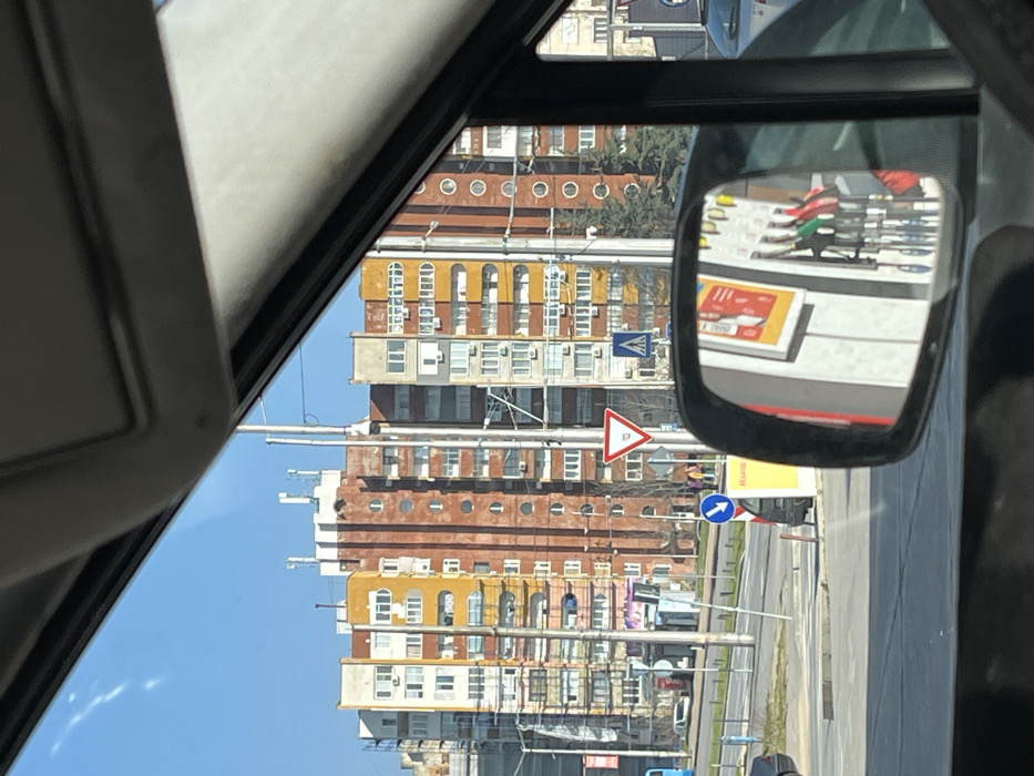 The image depicts an urban street scene in Sliven, Bulgaria, viewed from inside a stationary vehicle through the front windshield on a clear, sunny day. The sky above is bright blue with minimal cloud cover.
In the background, a row of multi-story residential apartment buildings dominates the skyline. These structures feature varied architectural styles and color palettes, including segments in faded yellow, brown, white, and grey. One prominent building in the center has a distinct brown facade with a vertical series of circular windows or decorative elements and is flanked by buildings with numerous rectangular windows and balconies. Many buildings display external air conditioning units and satellite dishes.
The midground features a wide asphalt road or intersection with multiple lanes, indicating a major thoroughfare. On the left side of the road, a blue public transit bus is partially visible, facing away from the viewer. Utility poles and streetlights line the road. Several traffic signs are clearly visible: a red and white triangular "Give Way" (Yield) sign, a blue square "Pedestrian Crossing" sign with a white figure, and a partially obscured blue circular sign with a white arrow. A marked pedestrian crosswalk is present on the asphalt. A small group of two to three individuals is observed walking on the sidewalk near the pedestrian crossing on the left.
The foreground is occupied by interior elements of the vehicle, specifically the upper portion of the windshield frame and the right-side rearview mirror. The mirror reflects a portion of what appears to be a Shell fuel station. Visible within the mirror's reflection are white structures, likely part of a pump island or canopy, a yellow sign with red graphics and text, and multiple fuel nozzles (with distinct red, green, black, and yellow handles) hanging from a dispenser unit. A grey vehicle is partially visible on the right side of the road, beyond the mirror.
