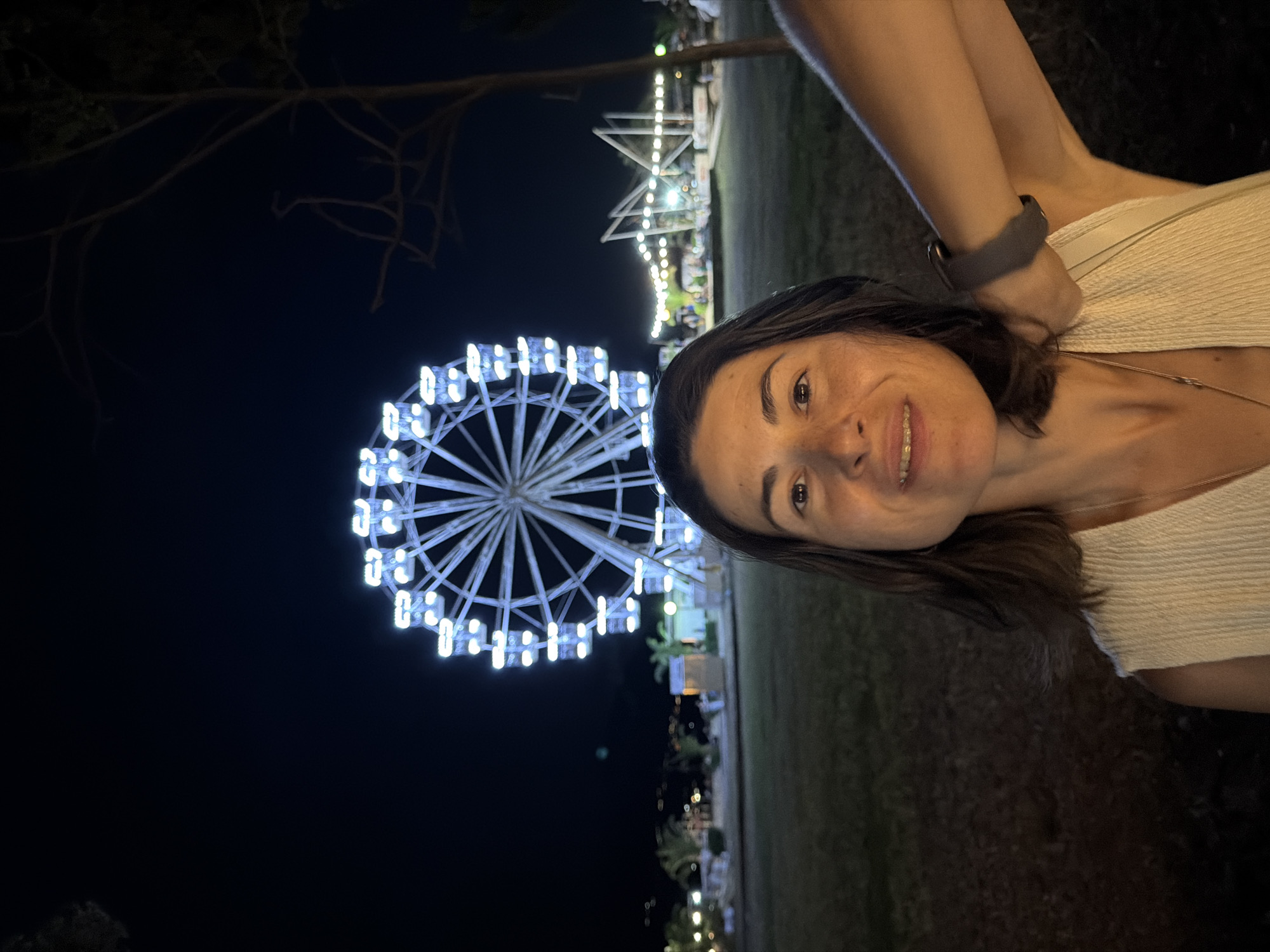 A woman smiles at the camera, her face in the foreground. She has dark hair, and is wearing a light-colored top. Behind her, a large Ferris wheel illuminated with white lights is visible against a dark night sky. The Ferris wheel is a prominent object in the scene. The setting appears to be an outdoor amusement park or fairground, with other lights and structures visible in the background, suggesting it is nighttime. The mood is cheerful, indicated by the woman's smile. There is no visible text in the image. The weather is not discernible, but the darkness suggests it is night. The location is unknown.