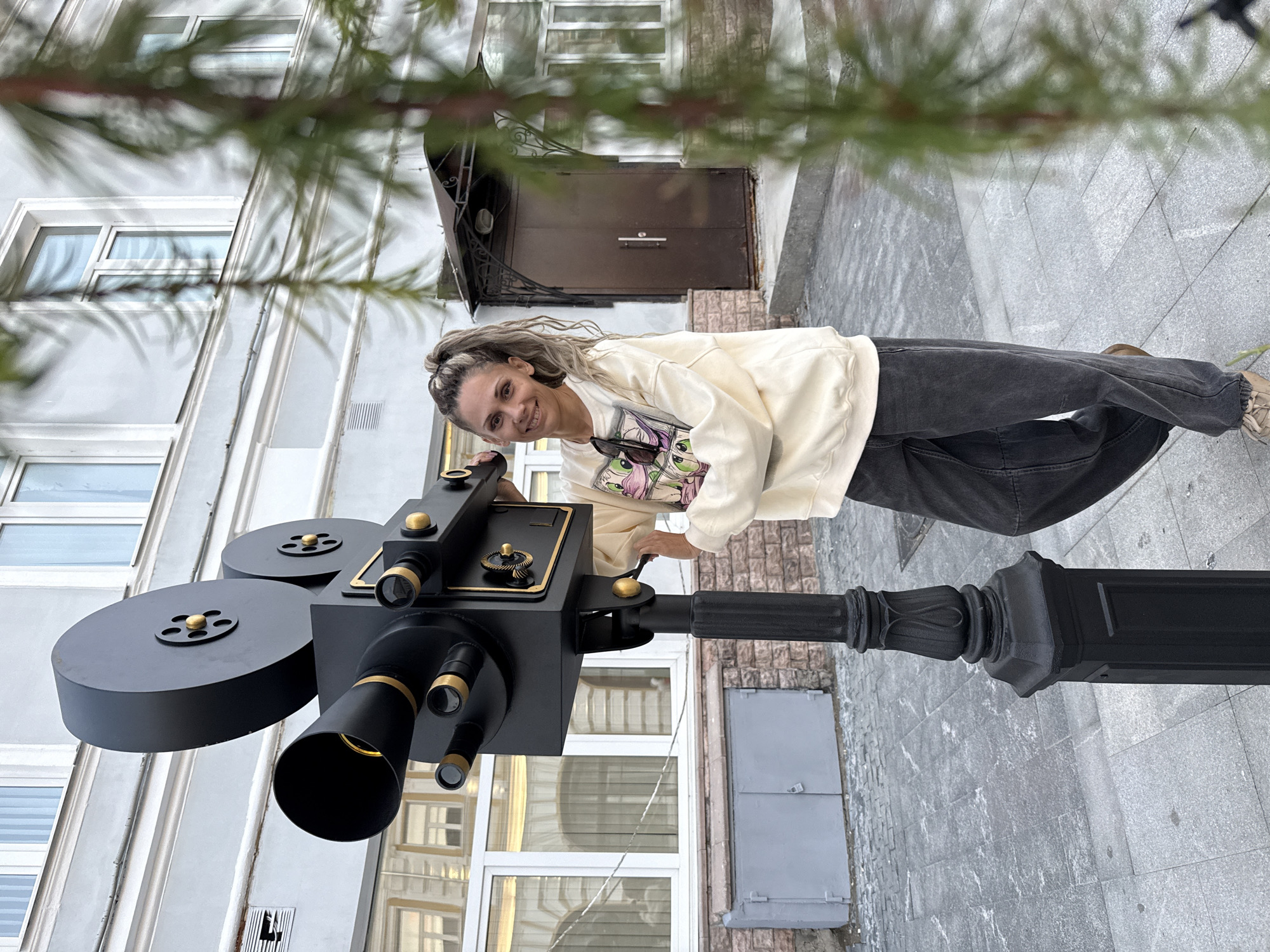A woman with blonde hair pulled into a ponytail stands next to a large, black, vintage-style movie camera prop mounted on a decorative black post. She is wearing a cream-colored sweatshirt with a graphic print and dark wash wide-leg jeans. The camera prop is detailed with gold accents and appears to be made of metal. The scene is set on a paved walkway in front of a building with light-colored walls and windows. Green foliage is visible in the foreground, blurring the background slightly. The background also shows a building entrance with a dark door and an awning, and more windows. The ground is paved with rectangular stones. The setting appears to be an outdoor urban environment, possibly a street or a courtyard. The location is identified as Rusanovo, Russia.