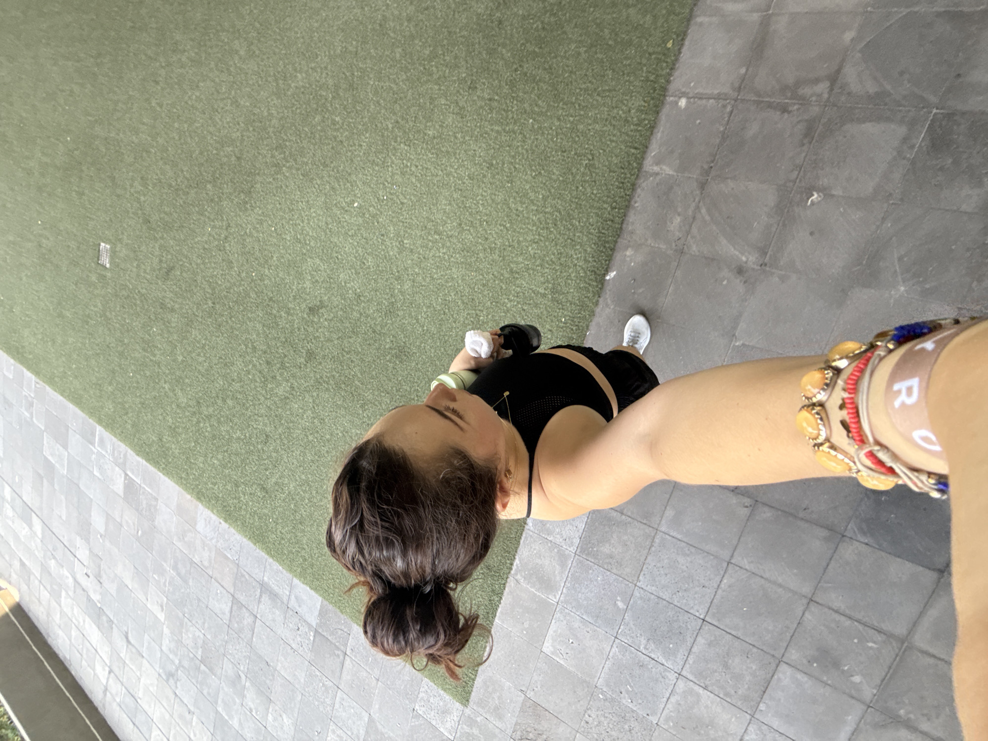 A woman takes a selfie while standing on a gray tile surface. The photo is taken from an elevated angle, looking down on the subject. She wears a black crop top and shorts, a white athletic shoe, and several bracelets on her right wrist. Her dark hair is tied in a bun. The background consists of artificial green turf on one side and gray tiles on the other, suggesting an outdoor urban environment. The location may be in Mexico City, Mexico.