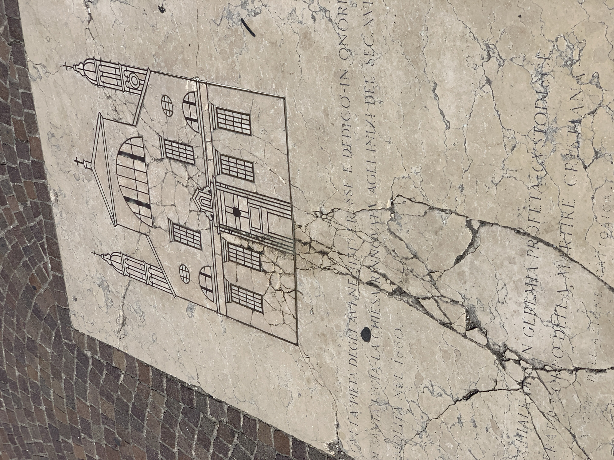 The image shows a cracked marble slab on a cobblestone ground. Engraved on the slab is an illustration of a church with two towers and a cross on top. Below the illustration, an inscription is partially visible, with some letters chipped or worn away. The scene is outdoors, with natural lighting. The cracks in the marble suggest age and wear. The cobblestone ground is laid in a patterned arrangement, and the overall impression is that of a historical marker or dedication. The location is in Venice, Italy, specifically on the facade of the Church of Santa Lucia.