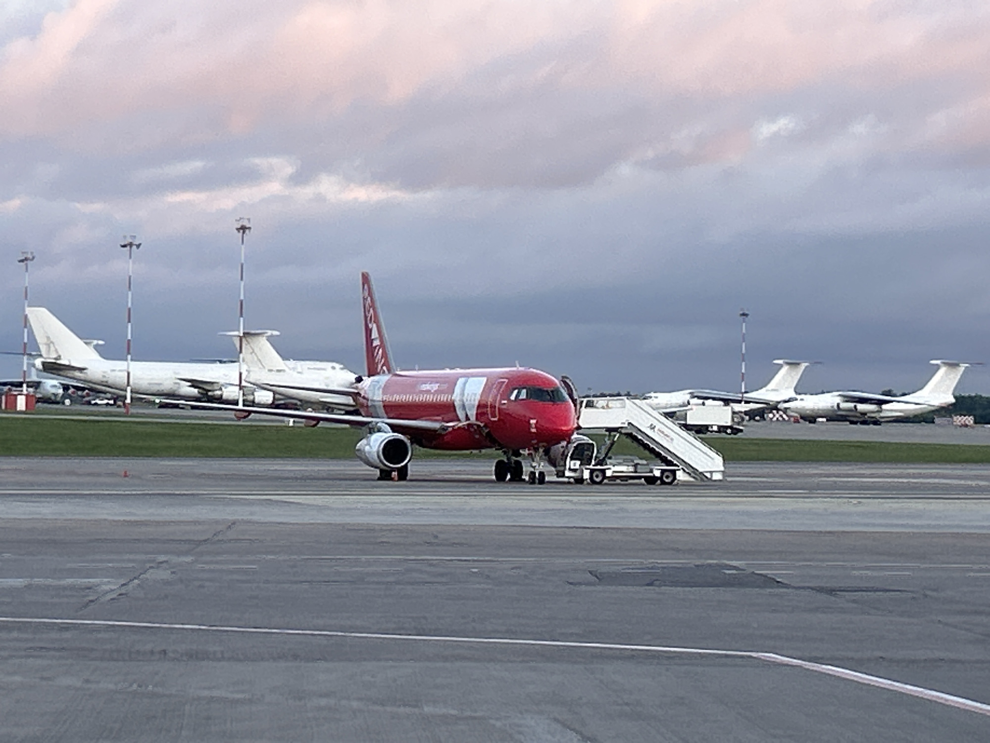 A red airplane with "RED WING" written on its tail is parked on an airport tarmac. A set of mobile stairs is connected to the airplane's door, and a small vehicle is parked nearby. In the background, several white airplanes are visible, along with tall poles with lights. The sky is cloudy with streaks of pink and purple from what appears to be either sunrise or sunset. The airport tarmac is gray and appears weathered in places. The scene is devoid of any visible people or discernible actions. The image captures a static view of an airport at dusk or dawn, likely in Minsk, Belarus, given the presence of Red Wings, which is a Russian airline that operates flights to Minsk.