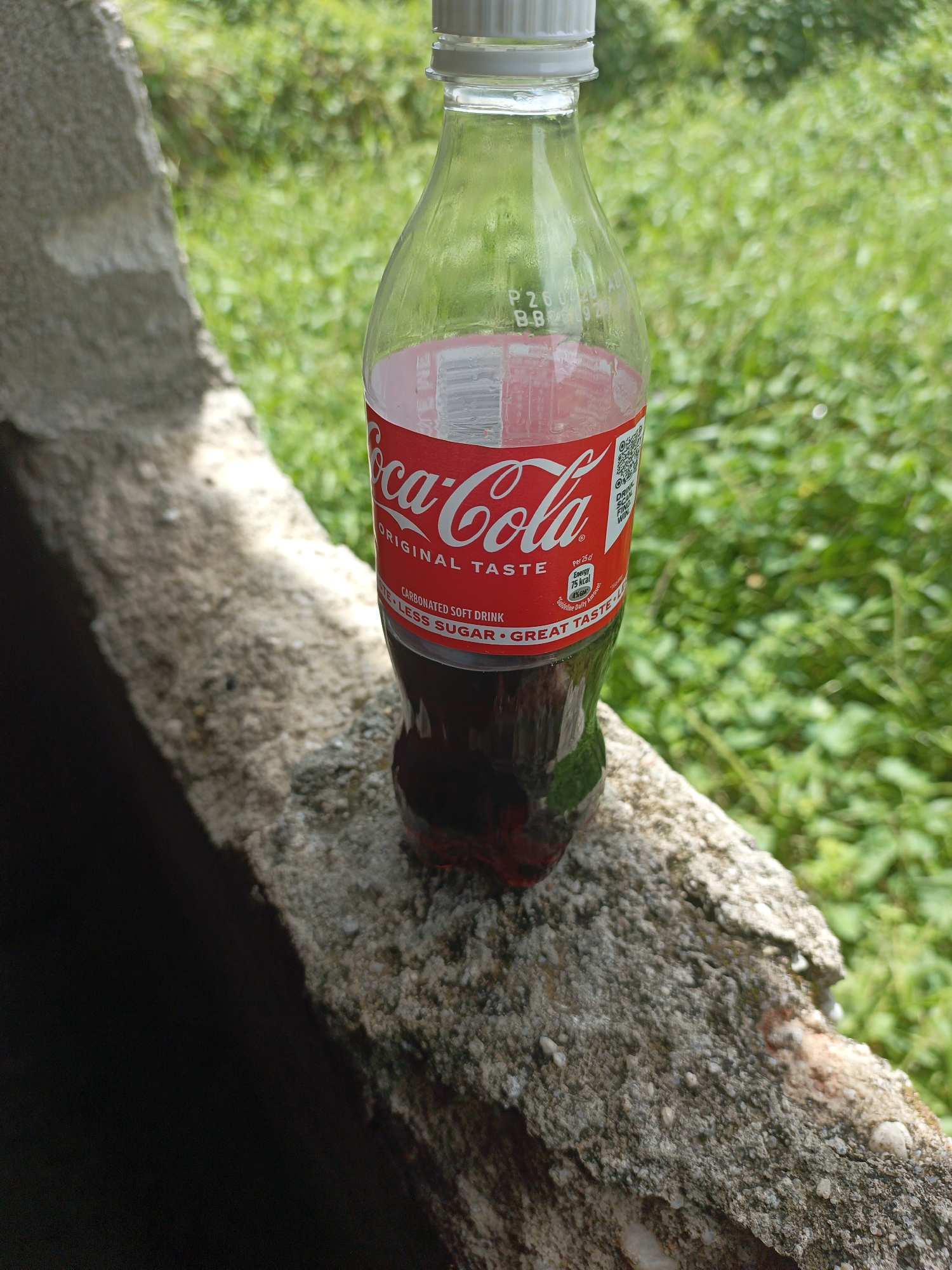 A partially full bottle of Coca-Cola stands on a weathered concrete surface. The bottle is plastic with a white cap, and the iconic Coca-Cola logo and label are visible. The background consists of green foliage, suggesting an outdoor setting. The Coca-Cola label shows that it is the original taste variety, but with less sugar. The OCR on the bottle reads "P2600led AL B-B920".