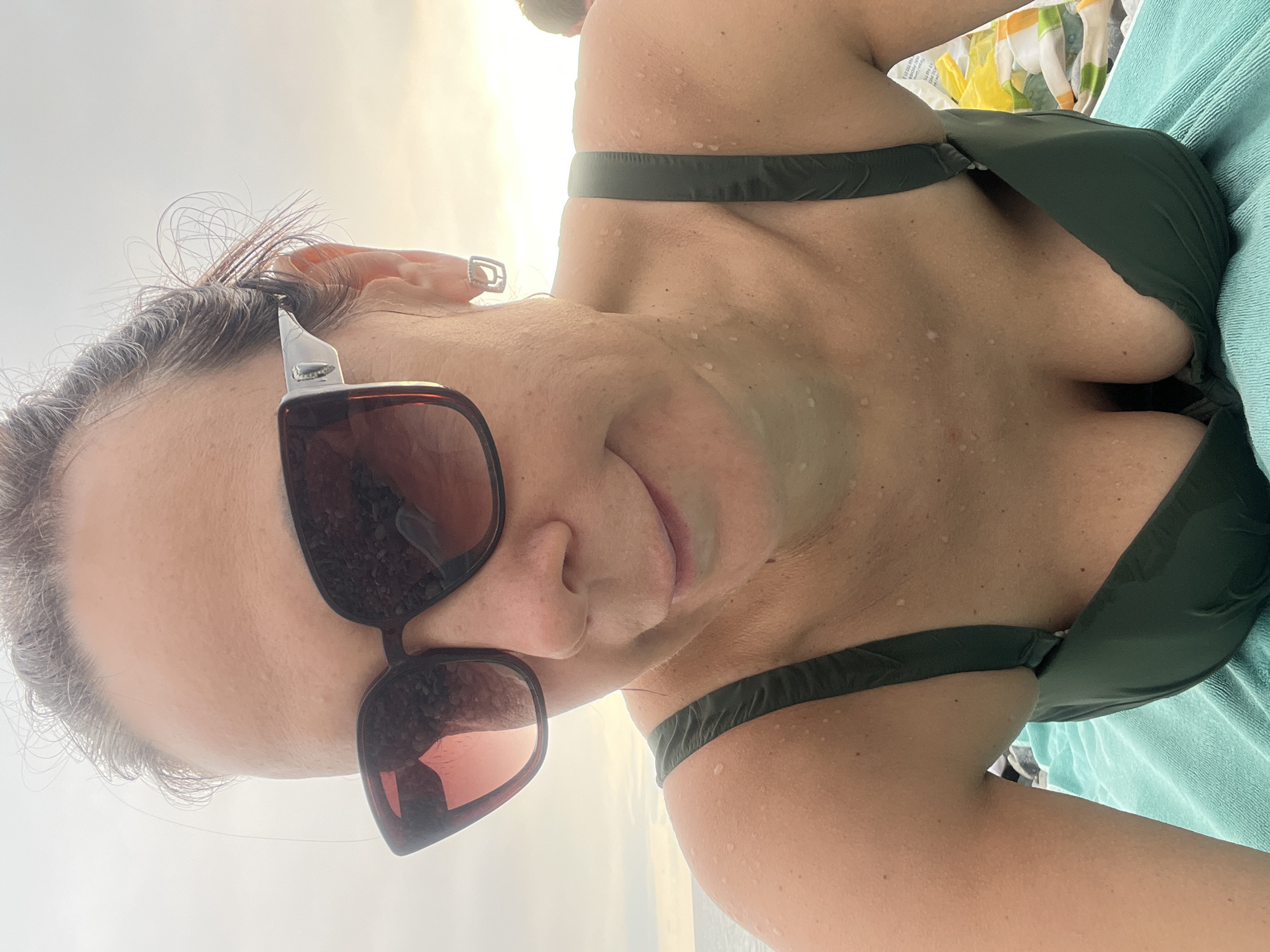 A woman with dark hair pulled back, wearing sunglasses and a green bikini, is captured in a selfie outdoors. Her skin appears dewy, suggesting she is near water. She is lying on a teal towel, with a beach or coastal environment visible in the background, characterized by a hazy sky and the suggestion of a body of water. The image is framed from the chest up, with her face angled downwards.