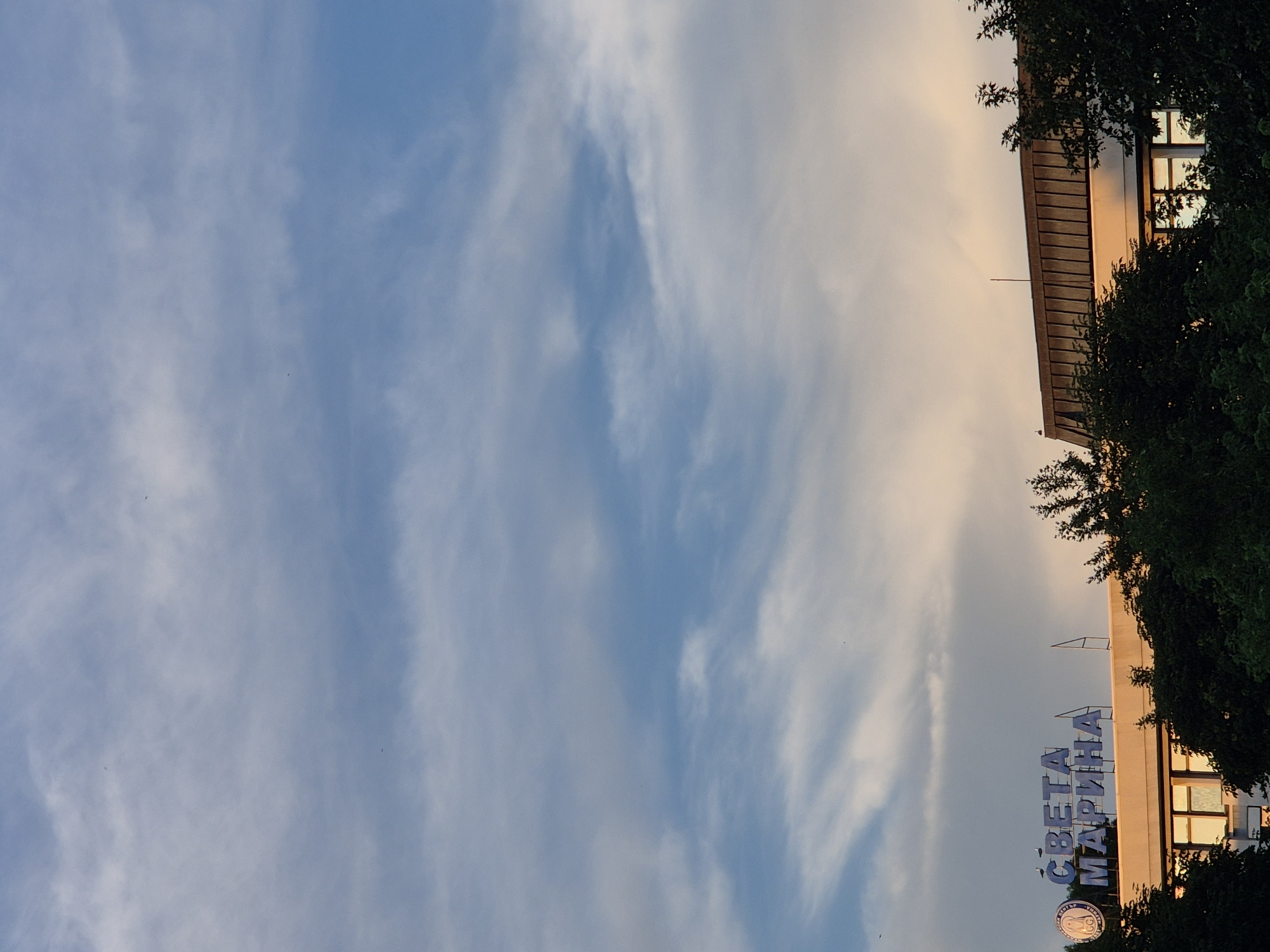 In a medium shot, a building's rooftop is visible under a sky with scattered clouds. The building has a sign with blue lettering that reads "СВЕТА МАРИНА" (Sveta Marina). The rooftop is flat and appears to be made of a dark material. Trees with dark green foliage surround the lower part of the building and extend into the foreground. The sky is a blend of blue and soft peach tones, suggesting it may be sunrise or sunset. There are no visible people or animals, but several small, dark specks in the sky could be birds. The overall impression is of an exterior urban scene during twilight.