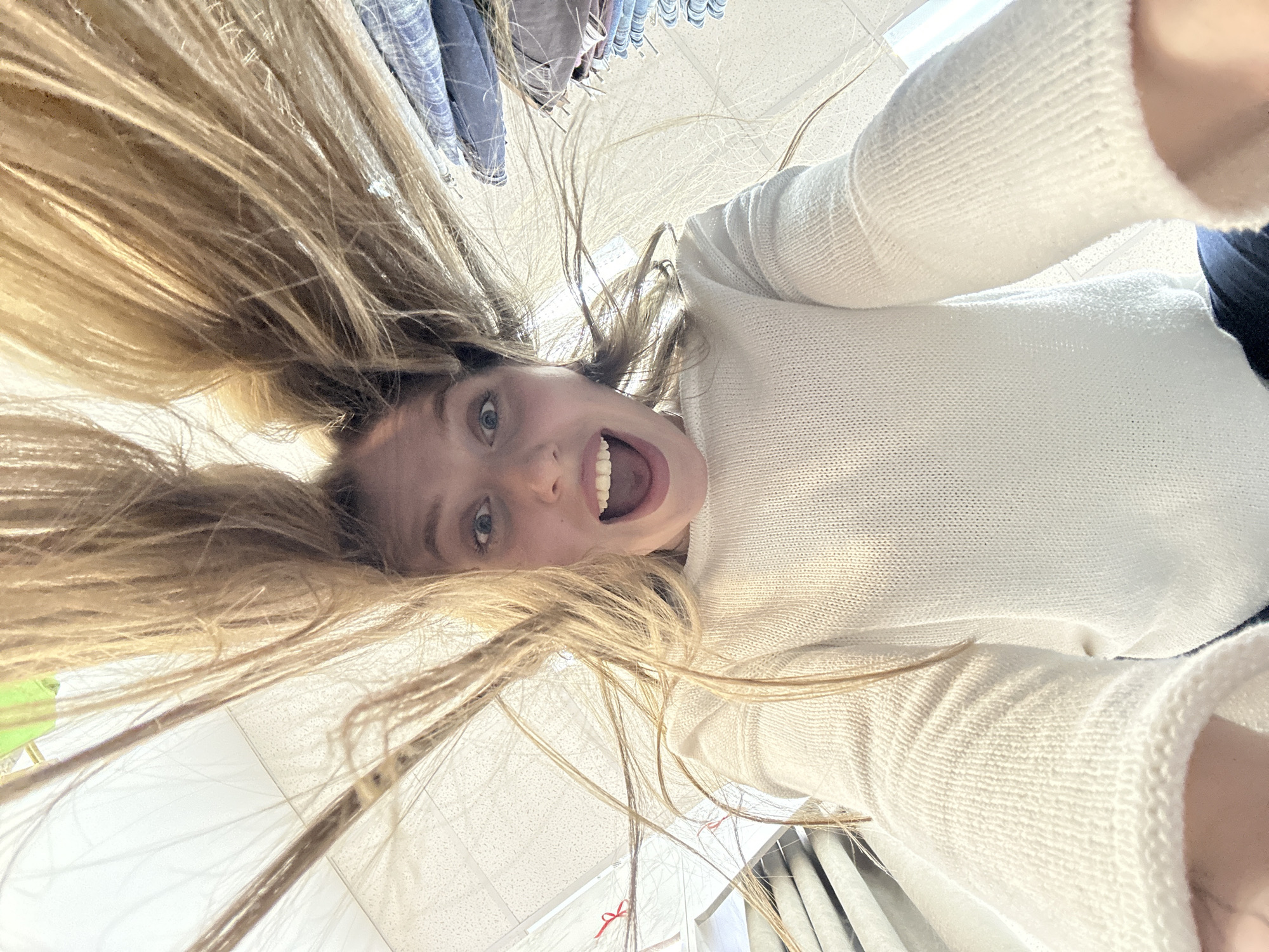 A person with long blonde hair is shown in a selfie-style image. The person, wearing a white sweater, is upside down, with their head towards the top of the frame and legs towards the bottom. Their mouth is open in a wide smile. The background appears to be a retail environment, possibly a clothing store, with clothes racks and ceiling tiles visible. The city of Chaikovsky, Russia, is not identifiable in the image.