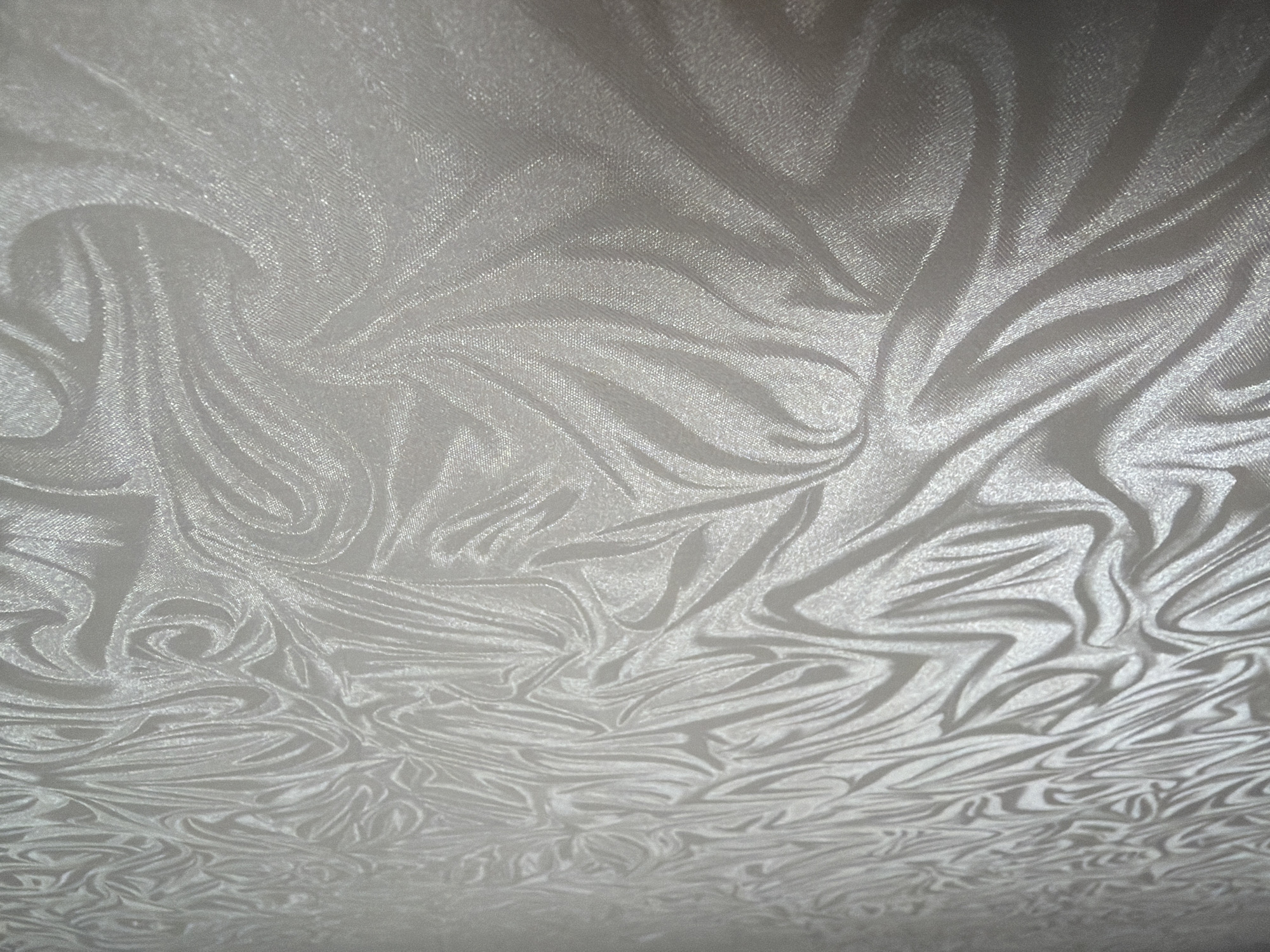 The image shows a close-up, low-angle view of a white, patterned fabric, possibly wallpaper or upholstery. The pattern consists of swirling, flowing lines and shapes that create a sense of movement and depth. The lighting highlights the texture of the fabric, with some areas appearing brighter and more reflective than others. There are no people or objects visible in the image, nor any environmental context to place it within Yuzhno-Sakhalinsk, Russia. The scene is entirely focused on the detailed texture and pattern of the material.
