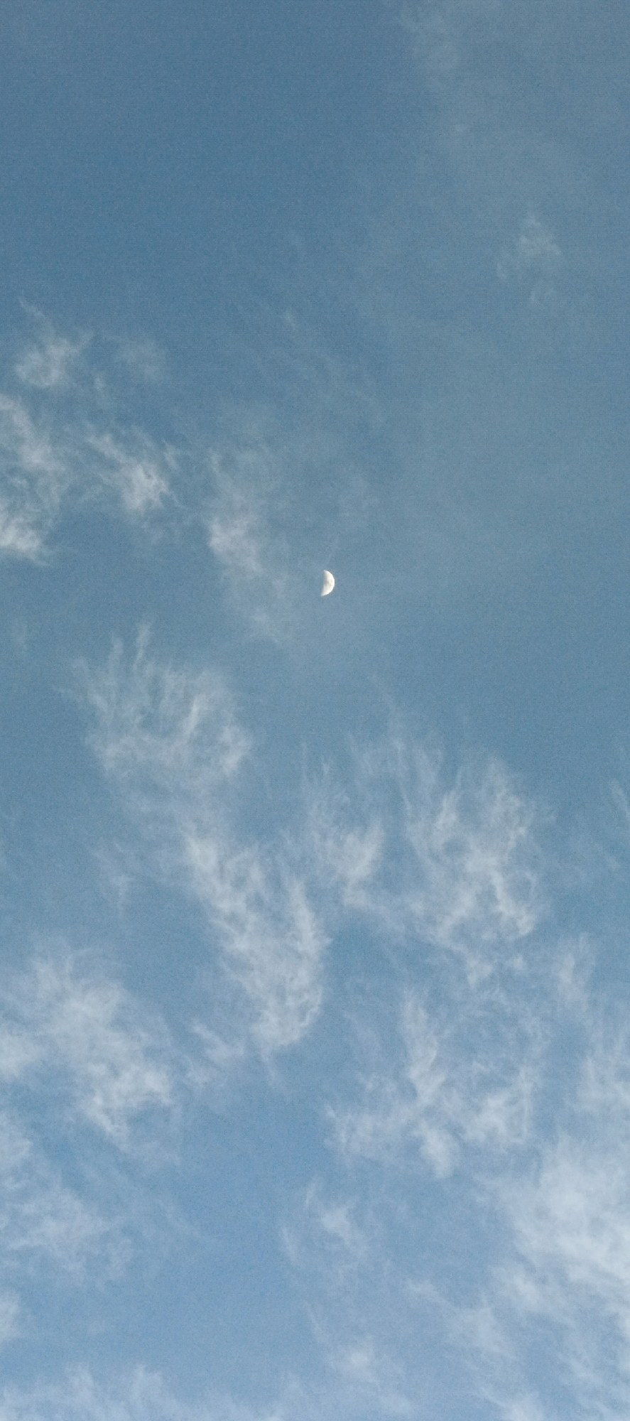 A crescent moon is visible in a clear, pale blue sky, partially obscured by wispy white clouds. The moon is located in the upper center portion of the frame. The clouds are scattered throughout the sky, creating a textured pattern. The image appears to be taken outdoors, likely during daylight or twilight. There are no people, objects, or discernible landmarks present in the image to suggest a specific location in Meunasahmesjid, Indonesia.