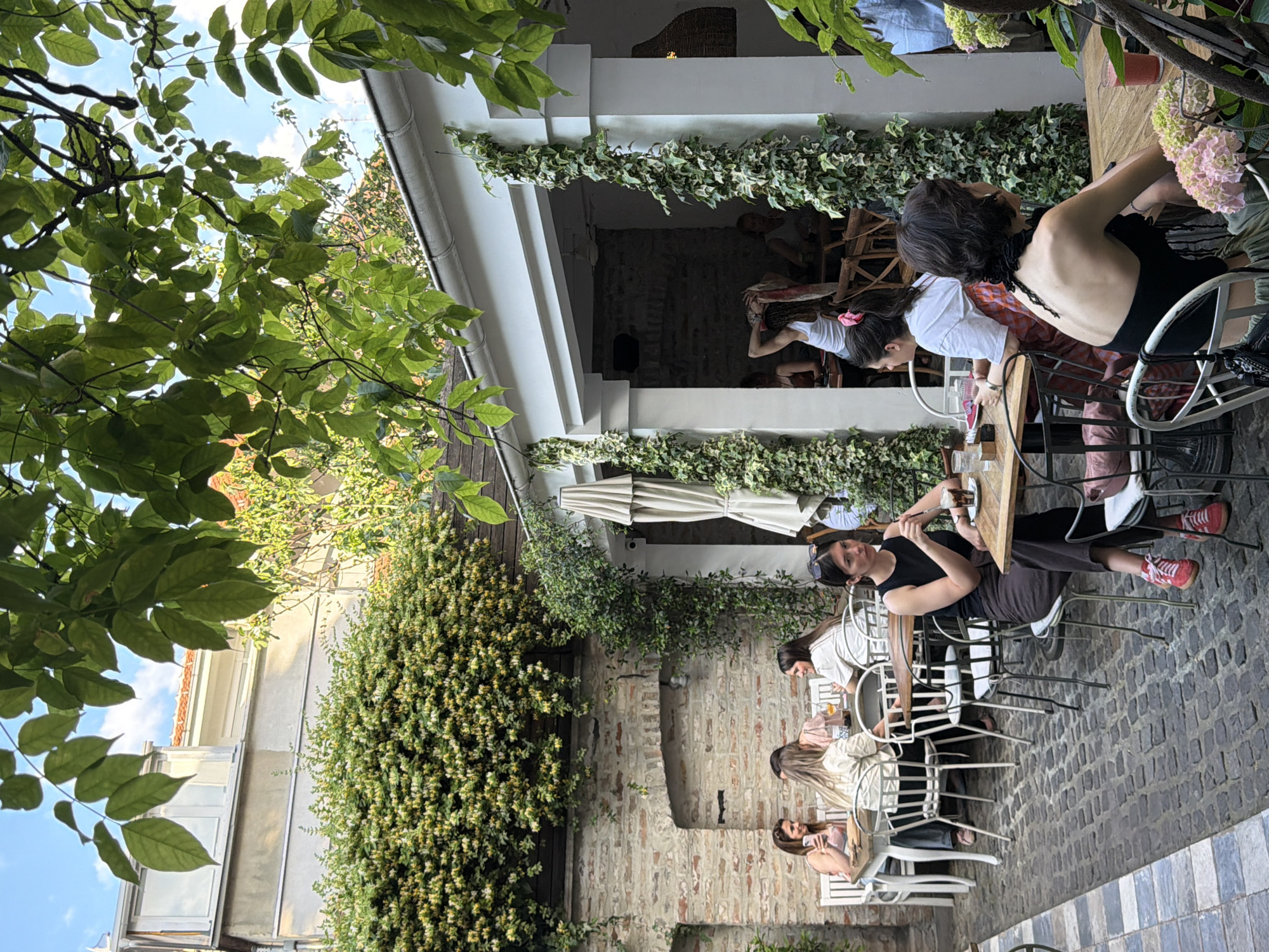 The image shows an outdoor courtyard cafe in Belgrade, Serbia. Several groups of people are seated at tables, dining and socializing. The courtyard has a brick floor and walls, with ivy and other greenery covering the walls and overhead structures. The cafe features white columns and archways. Some people are visible inside the building. A mix of modern and traditional architecture is present. The atmosphere appears relaxed and casual. The sky is visible through the leaves of a tree.