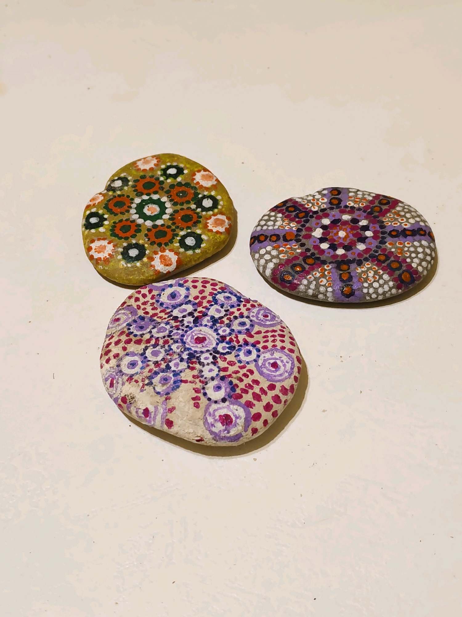 Three painted rocks are arranged on a light-colored, textured surface. The rocks are decorated with intricate dot patterns in various colors, including green, orange, white, black, purple, and pink. One rock is predominantly green and orange, another is purple and orange, and the third is a mix of pink and purple. The background appears to be a table or countertop. There is no information in the image to suggest a location in Rostov-on-Don, Russia.