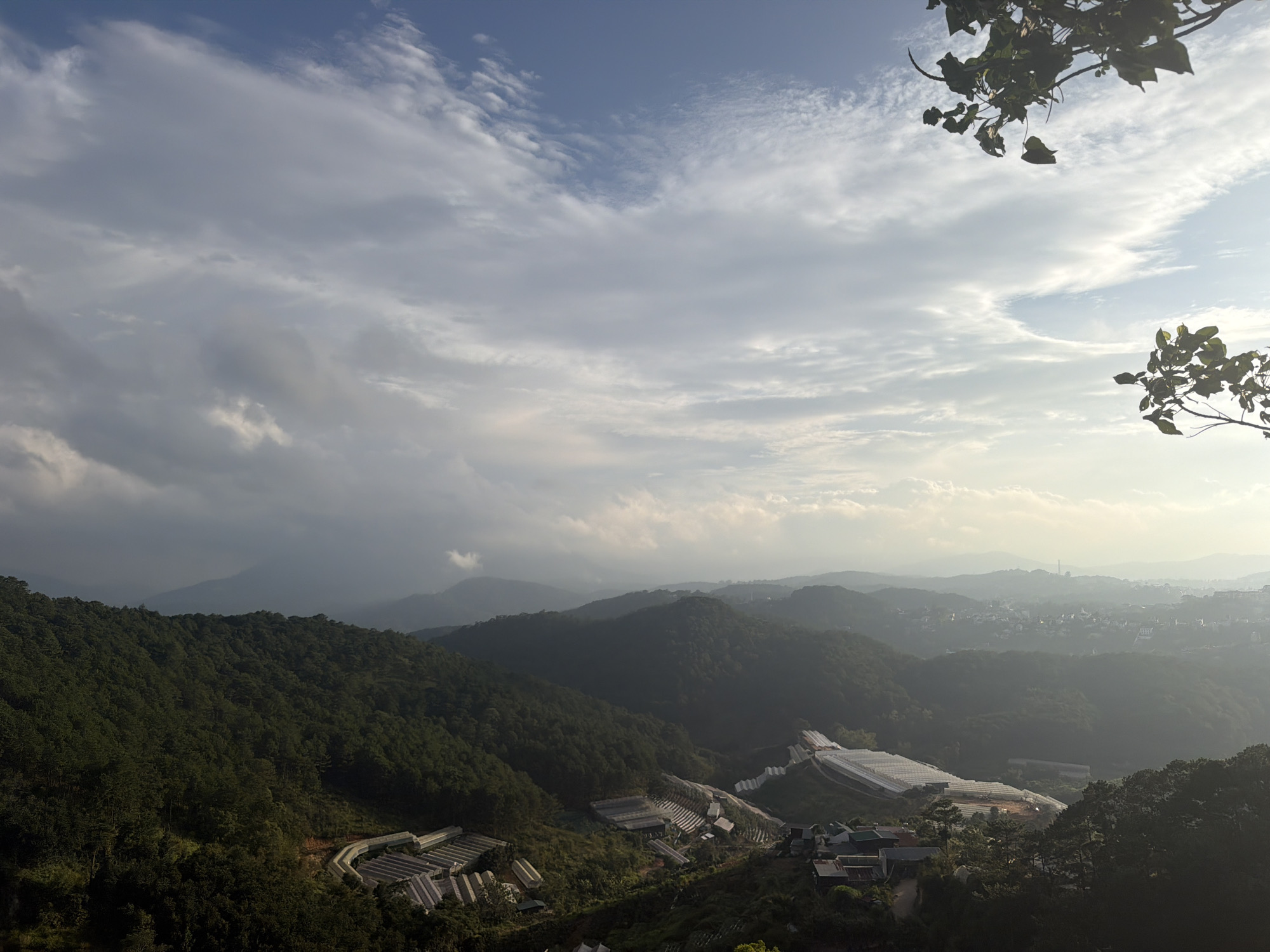 The image depicts a panoramic view of a mountainous region with agricultural greenhouses and a distant town. The sky is filled with scattered clouds, and sunlight filters through, creating a hazy atmosphere. In the foreground, lush green trees cover the hillsides. Several rows of white greenhouses are visible, suggesting an agricultural area. In the mid-ground, the landscape transitions into rolling hills that recede into the distance. A town with buildings can be seen nestled among the hills in the background. The image appears to be taken from a high vantage point, offering a wide vista of the natural and developed landscape. The location is identified as Xuân Thành, Vietnam. There are no people or animals clearly visible in the frame.