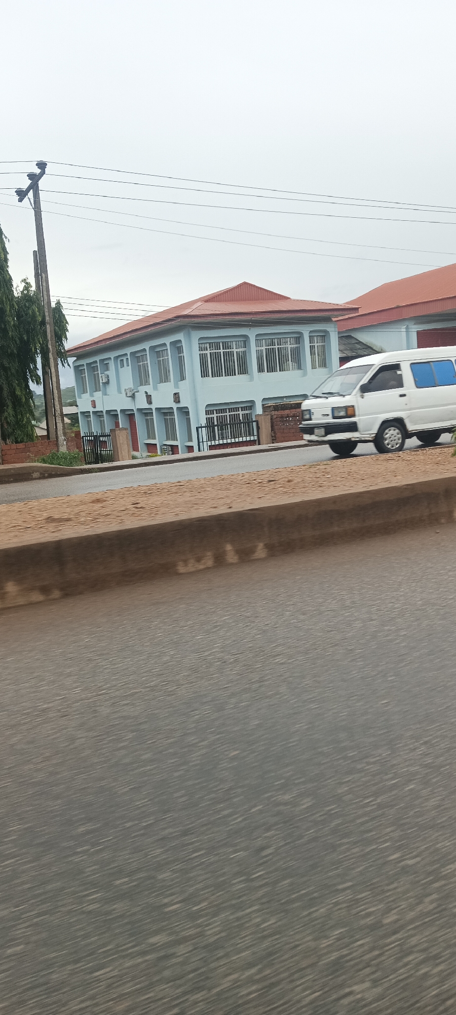 The image captures a street scene in Bukuru, Nigeria, under an overcast sky during daytime. The prominent subject is a two-story building on the right side of the frame, featuring light blue walls and a red, corrugated metal roof. The building has numerous windows, many of which are fitted with white vertical security bars. Several air conditioning units are visible on its exterior. A black metal fence runs along the front of the property, which is bordered by a concrete curb and a patch of reddish-brown earth.
To the right of the building, a white van, possibly a Toyota Hiace or similar model, is either moving or parked on the paved road. The van has blue-tinted rear windows. A dark wooden utility pole with multiple horizontal cross-arms and overhead wires stands on the left side of the frame, partially obscured by green foliage from a tree. The foreground consists of a grey asphalt road, displaying some motion blur, suggesting the photo was taken from a moving vehicle. No discernible text is visible on any of the subjects.