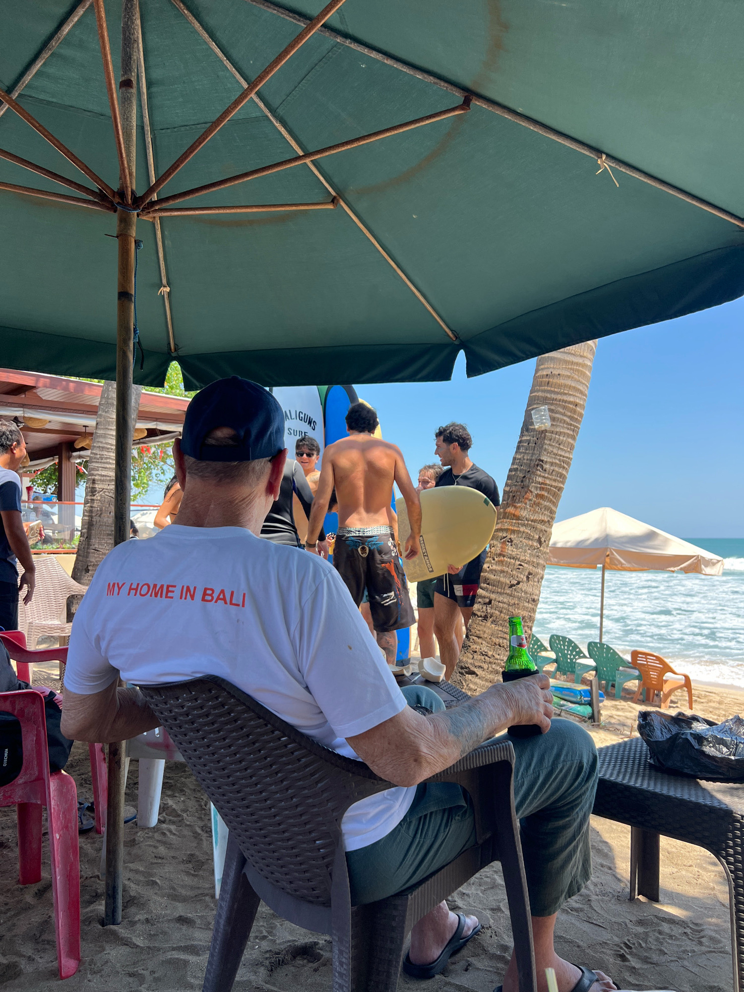 The image depicts a sunny beach scene in Kuta, Indonesia, likely a popular tourist spot given the presence of surfboards and beach umbrellas. In the foreground, an older man wearing a white t-shirt with "MY HOME IN BALI" printed on it in orange, and a dark blue baseball cap, is seated in a brown plastic chair, holding a green bottle of beer. He is facing away from the camera, towards the ocean. His legs are clad in olive green pants, and he wears black flip-flops.
Behind him, several individuals are gathered, some holding surfboards. One man in the middle ground, with a shirtless torso, appears to be talking to another man holding a light yellow surfboard. They are standing near a palm tree. A few beach chairs, some empty and some occupied, are visible, as are some surfboards leaning against a building in the background. The sand is light brown, and the ocean is a clear blue under a bright sky. The overall atmosphere suggests a relaxed, casual day at the beach.