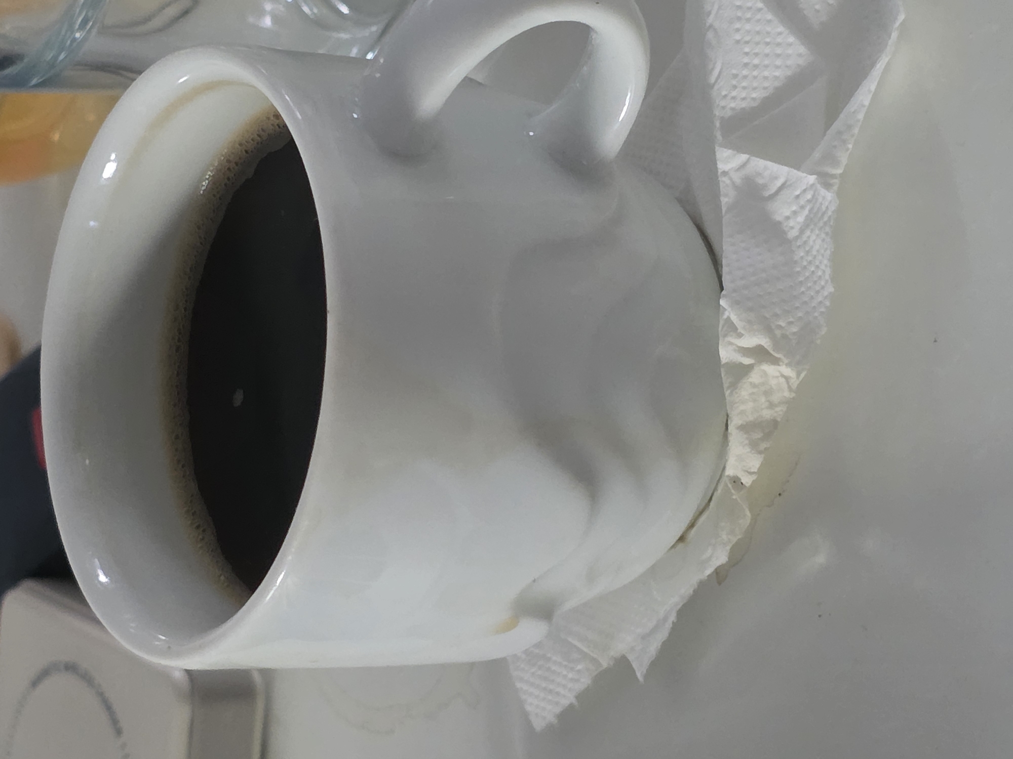 A white ceramic mug, filled with dark coffee, sits on a white surface, possibly a table or tray. The mug has a slightly irregular, wavy texture on its exterior. A white paper napkin is folded beneath the mug, with its edges slightly fanned out. In the background, a portion of a light-colored object, possibly a device or another container, is visible to the left. To the right, a clear glass container with an orange liquid can be partially seen. The scene appears to be indoors, with soft lighting. The location is identified as Unknown, Unknown.