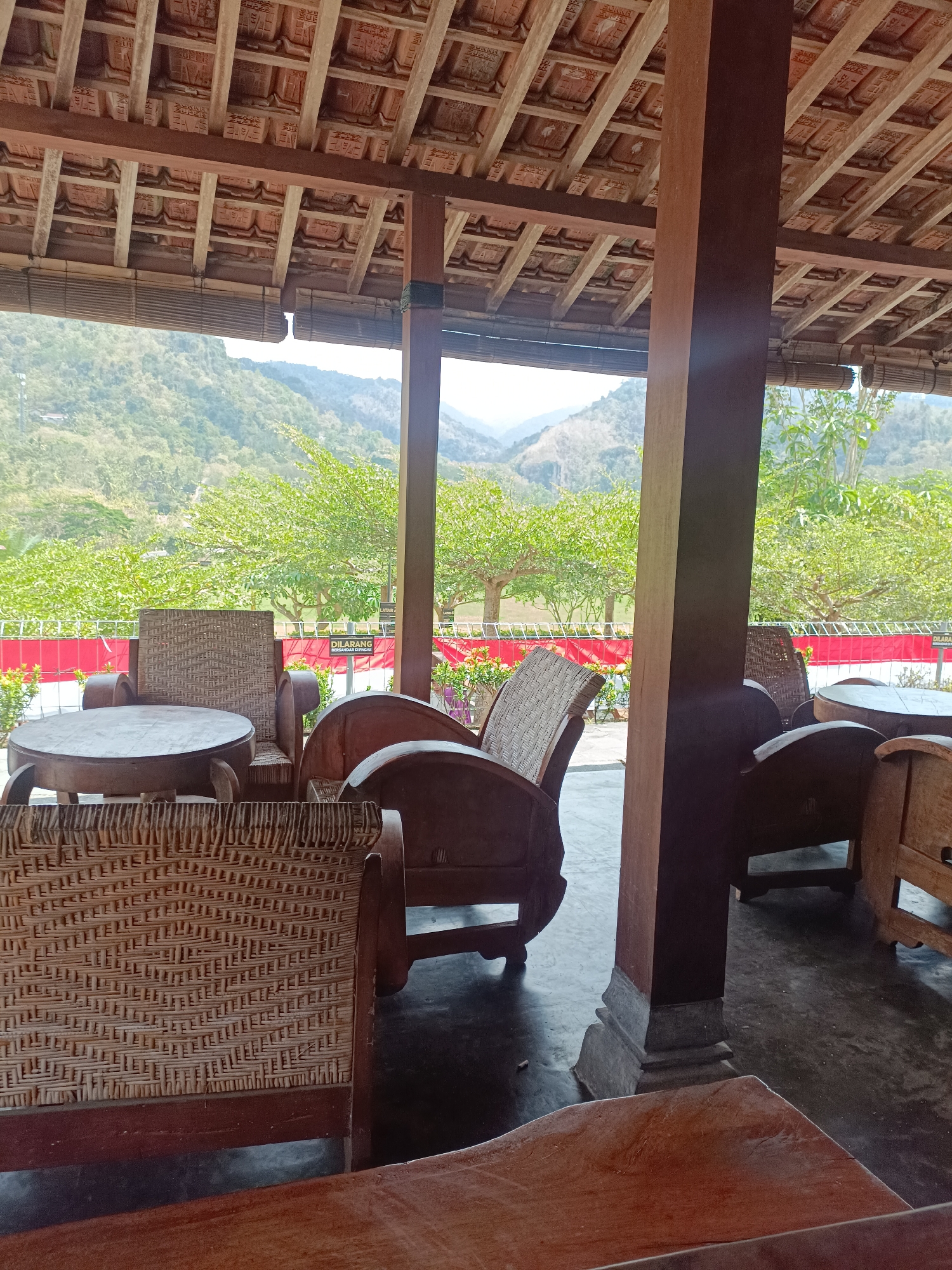 This image depicts a seating area with wooden furniture and a thatched roof, set against a backdrop of lush green hills and trees, likely in Pendoworejo, Indonesia. The scene appears to be an outdoor or semi-outdoor space, possibly a cafe or a traditional rest area. Several woven chairs and tables are visible, arranged under a brown wooden support beam and a tiled roof. In the background, a red and white striped fence separates the seating area from a natural landscape. The overall atmosphere is tranquil and inviting, suggesting a place to relax and enjoy the scenic view.