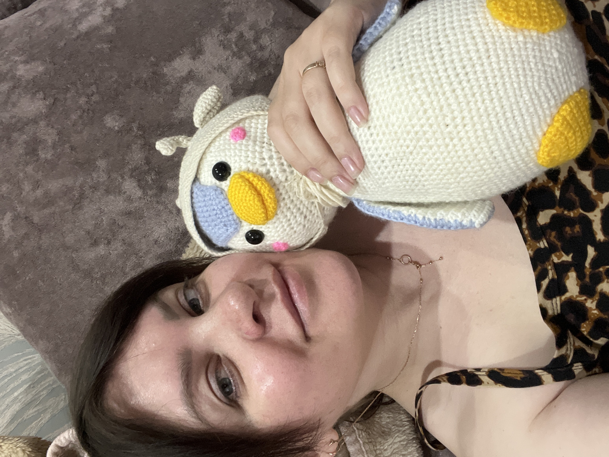 A woman lies on a brown textured surface, possibly a couch, with a crocheted owl toy resting on her chest. She gazes upwards, slightly to her left. The woman has dark hair and light skin. She wears a gold necklace. Her right hand is holding the owl, and a gold ring is visible on her ring finger. She is wearing a leopard-print top. The location of the scene is Noviy, Russia.