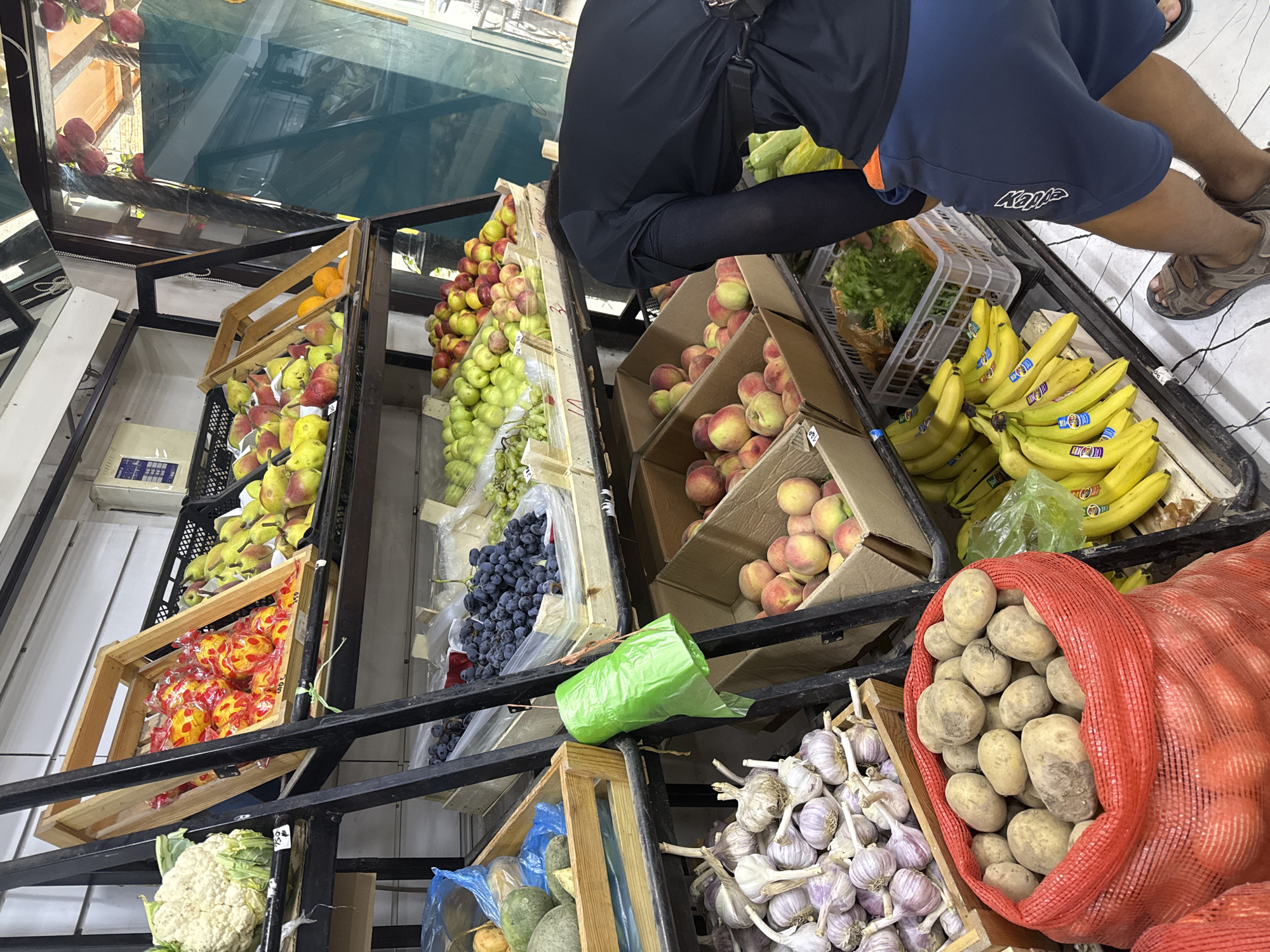 The image depicts a produce stall, likely in Osh City, Kyrgyzstan, displaying a variety of fruits and vegetables. In the foreground, a large red mesh bag is filled with potatoes. To the left, several bulbs of garlic are arranged in a wooden crate. Above the garlic, there are avocados in another wooden crate, and further up, cauliflower.
The middle section of the stall features shelves with different fruits. One shelf holds grapes in plastic bags, with other fruits like apples and pears visible in wooden crates. Another shelf displays peaches in cardboard boxes. A person wearing a black t-shirt and blue shorts is bending over, appearing to interact with the produce.
The background shows more shelves with fruits, including what looks like citrus in wooden crates and bananas in a crate. There are also bundles of bananas hanging. The stall has a rustic appearance with wooden crates and metal shelving. The lighting suggests it is daytime, possibly indoors or under a covered area.