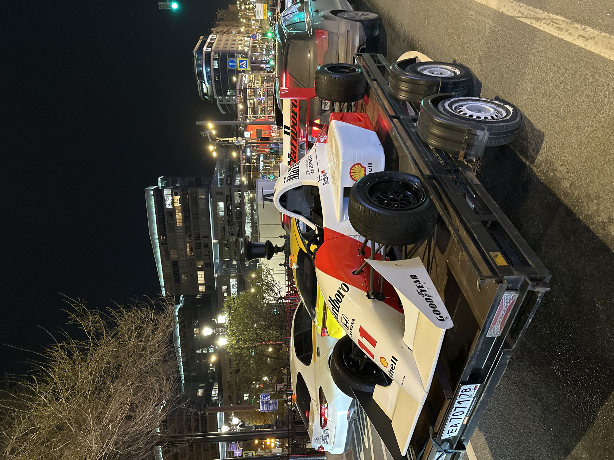 Here's a matter-of-fact description of the image:
The image shows a Formula 1 race car, specifically a McLaren-Honda with Marlboro livery and number "11", loaded on a trailer. There are spare tires also on the trailer. The trailer has license plate "EA 707178" with a "RUS" indicator, suggesting it's registered in Russia.
In the background, there's an urban landscape at night with buildings, street lights, and traffic signals. A car with license plate "K939AE977" is also visible.