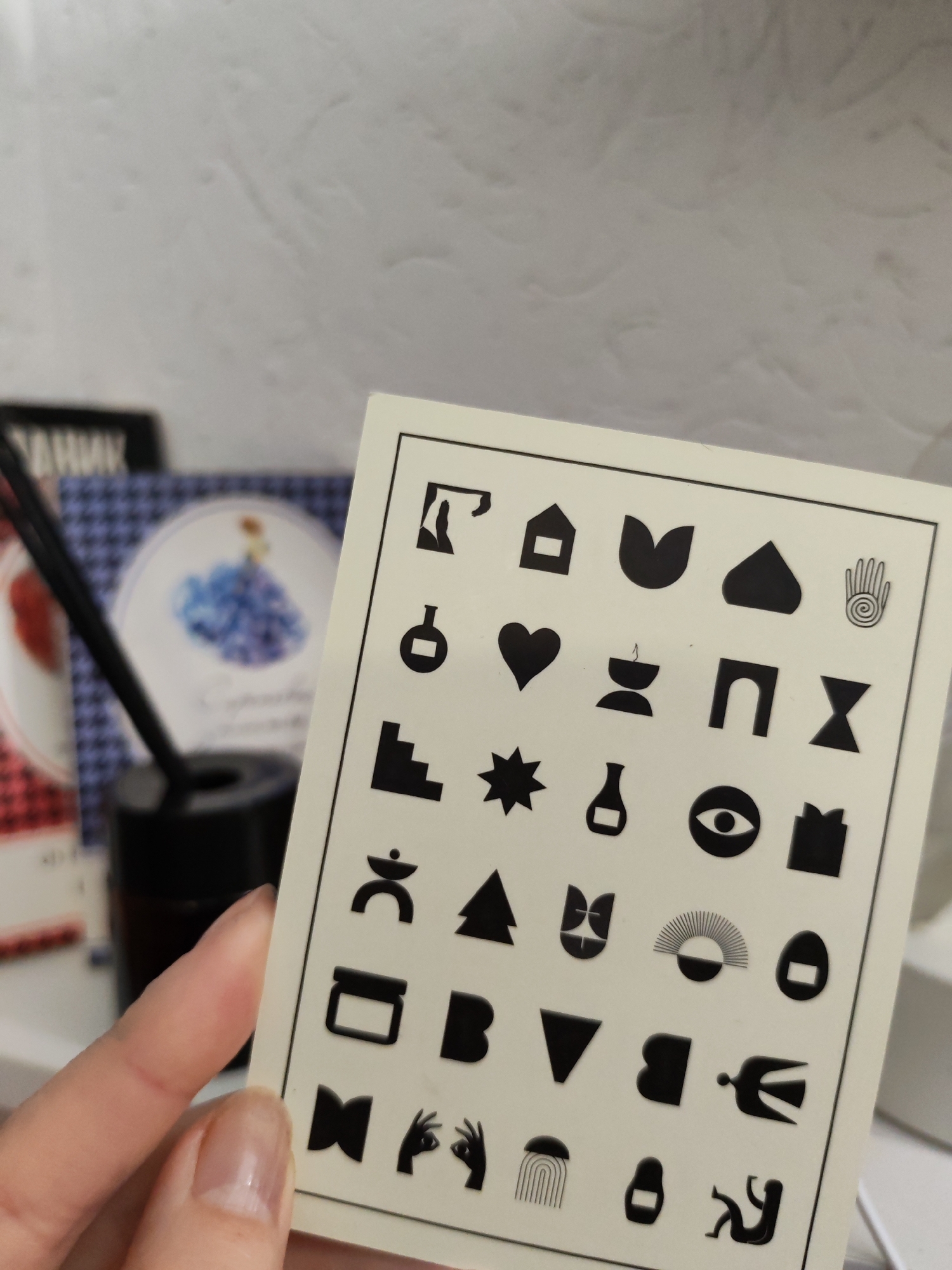 The image shows a hand holding a white card with black minimalist symbols arranged in a grid. The symbols include a house, a tulip, a heart, an eye, a bottle, and a hand. The background includes a white wall, a diffuser with black sticks in a black container, and a book with a flower illustration on the cover. There is no information in the image to confirm the location as Izhevsk, Russia.