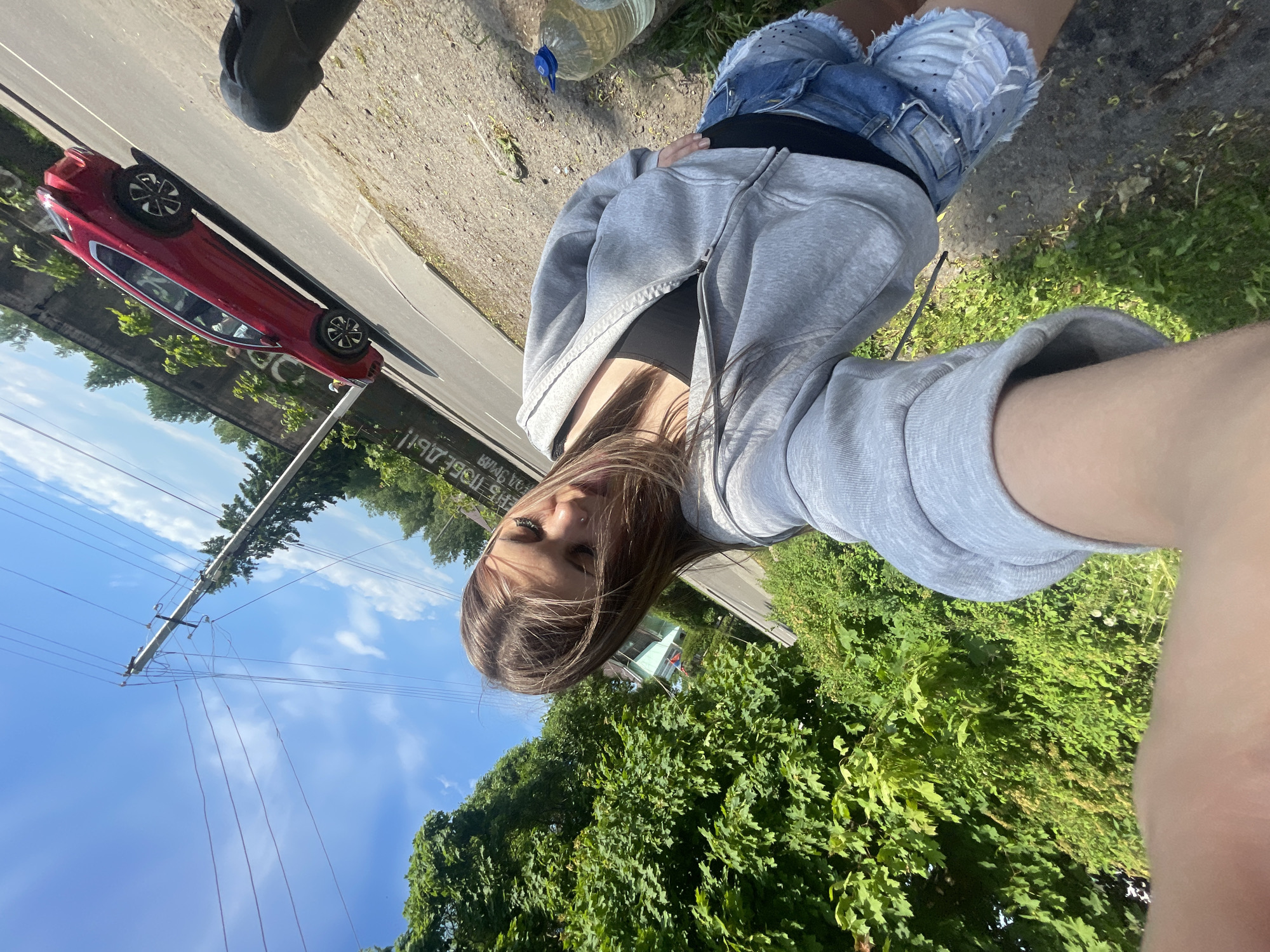 The image depicts an outdoor selfie taken in Priozersk, Russia, under bright daylight conditions. The frame is significantly tilted.
In the immediate foreground, a young woman is visible, appearing to hold the camera. Her long, light brown hair, partially obscures her face, and a nose piercing is present on her right nostril. She is wearing a grey zip-up hoodie over a black top and light blue distressed denim shorts. Her right forearm and hand, which hold the camera, are visible in the lower left portion of the image.
The background shows a paved road, sloping diagonally from the lower left to the upper right. A red compact crossover SUV is visible on the right side of the road, oriented with its rear towards the viewer. Adjacent to the road, on the right, is a dirt or gravel verge where a large, clear plastic water bottle with a blue cap rests. Behind the road, a concrete or stone retaining wall with visible Cyrillic graffiti, including a partially legible word like "ПОВЕДЬ!", forms part of the distant background.
To the left of the woman, dense green foliage from numerous trees and bushes occupies a significant portion of the frame. A light-colored building with a roof is partially discernible through the trees on the far left. Above the landscape, a tall utility pole with multiple power lines extends across a clear blue sky, which contains scattered white cumulus clouds. The environment suggests a sunny, temperate day with abundant vegetation.