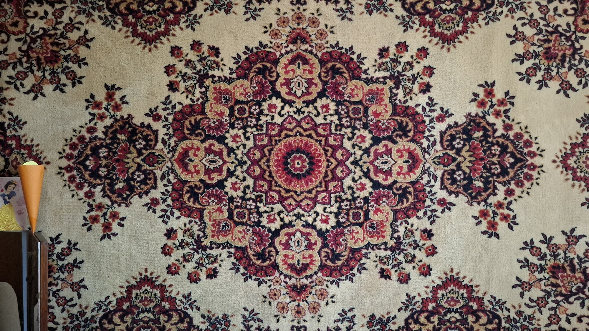 The image shows a detailed view of a cream-colored carpet with a floral and geometric pattern. The dominant colors in the pattern are deep red, burgundy, black, and beige. The central motif is a large, intricate mandala-like design. Scattered throughout the carpet are smaller floral clusters and repeating patterns. On the left side of the image, a portion of a wooden shelf is visible, supporting an orange cone-shaped object and a small framed picture of a woman in a yellow dress. The carpet appears to be hanging on a wall or draped over a surface. The setting is indoors, with the lighting suggesting a well-lit room. No specific location in the city of Shymkent, Kazakhstan can be identified from the visual information provided.