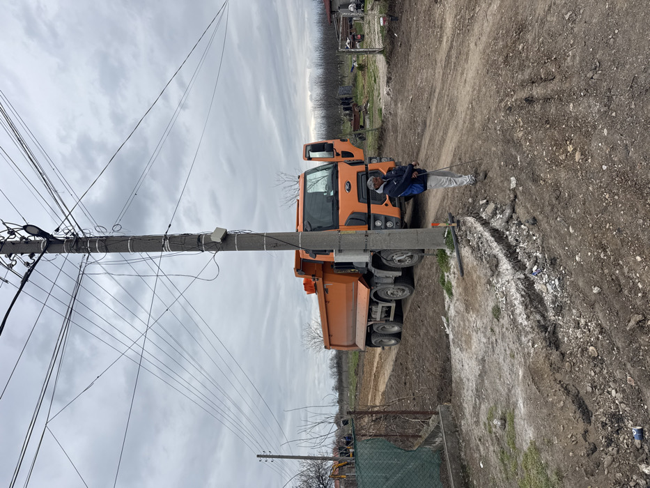 The image depicts an outdoor scene in Kochevo, Bulgaria, under an overcast sky. A large, orange, multi-axle Ford dump truck is parked on a dirt and gravel road, positioned towards the center-right, with its driver's side door open. In the foreground, slightly to the right of the truck and centrally placed, stands a male individual with gray hair, wearing a dark blue and light blue jacket, light-colored pants, and dark shoes. He faces right, holding a slender metal rod or stick diagonally downwards in both hands.
Immediately to the man's left and extending into the foreground is a concrete utility pole supporting numerous interwoven power and communication lines. A street light fixture and a small white rectangular device are attached to the pole. At the base of the pole, a small orange marker is visible. The dirt road in the immediate foreground shows signs of recent excavation, with a trench-like depression and disturbed earth. A blue aluminum can lies on the ground to the left of this trench.
To the left of the road, a green chain-link fence borders what appears to be a cleared or cultivated area, beyond which a yellow excavator is visible in the distance. To the right of the road, another fence and a metal gate separate the road from a more open, unkempt area. The background features numerous bare trees, consistent with an orchard or rural landscape, and indistinct figures are scattered in the mid-ground and background.