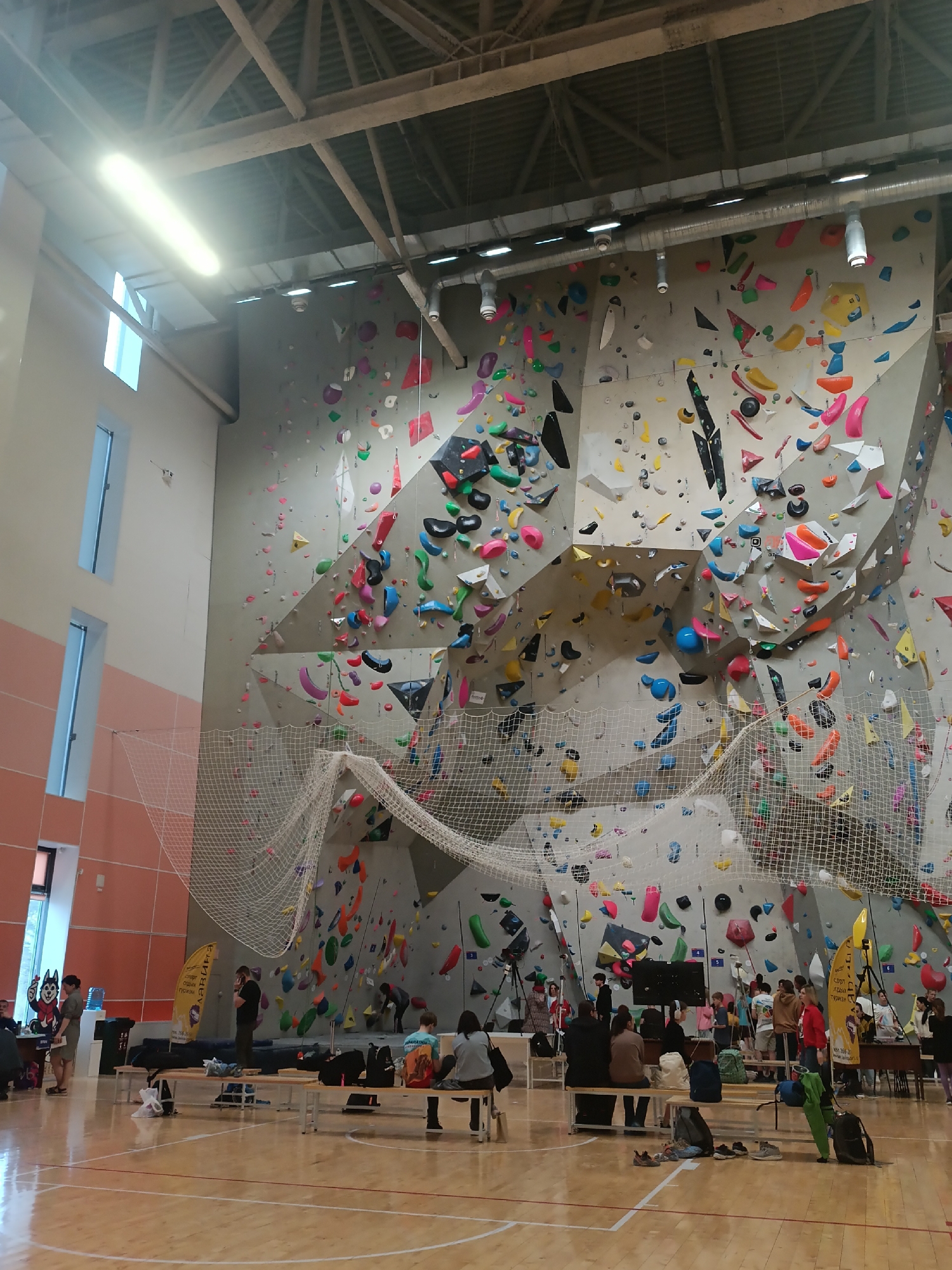 The image depicts the interior of a climbing gym in Krasnoyarsk, Russia. A large climbing wall with multicolored holds dominates the background. A safety net is stretched across the lower section of the wall. In the foreground, people are sitting on benches. Some people stand nearby. Banners are visible on the left and right of the seating area. The gym has a high ceiling with visible support beams and lighting. The floor is made of wood and shows basketball court markings. Windows are visible on the left. A person in a wolf costume is present near the left edge of the frame.