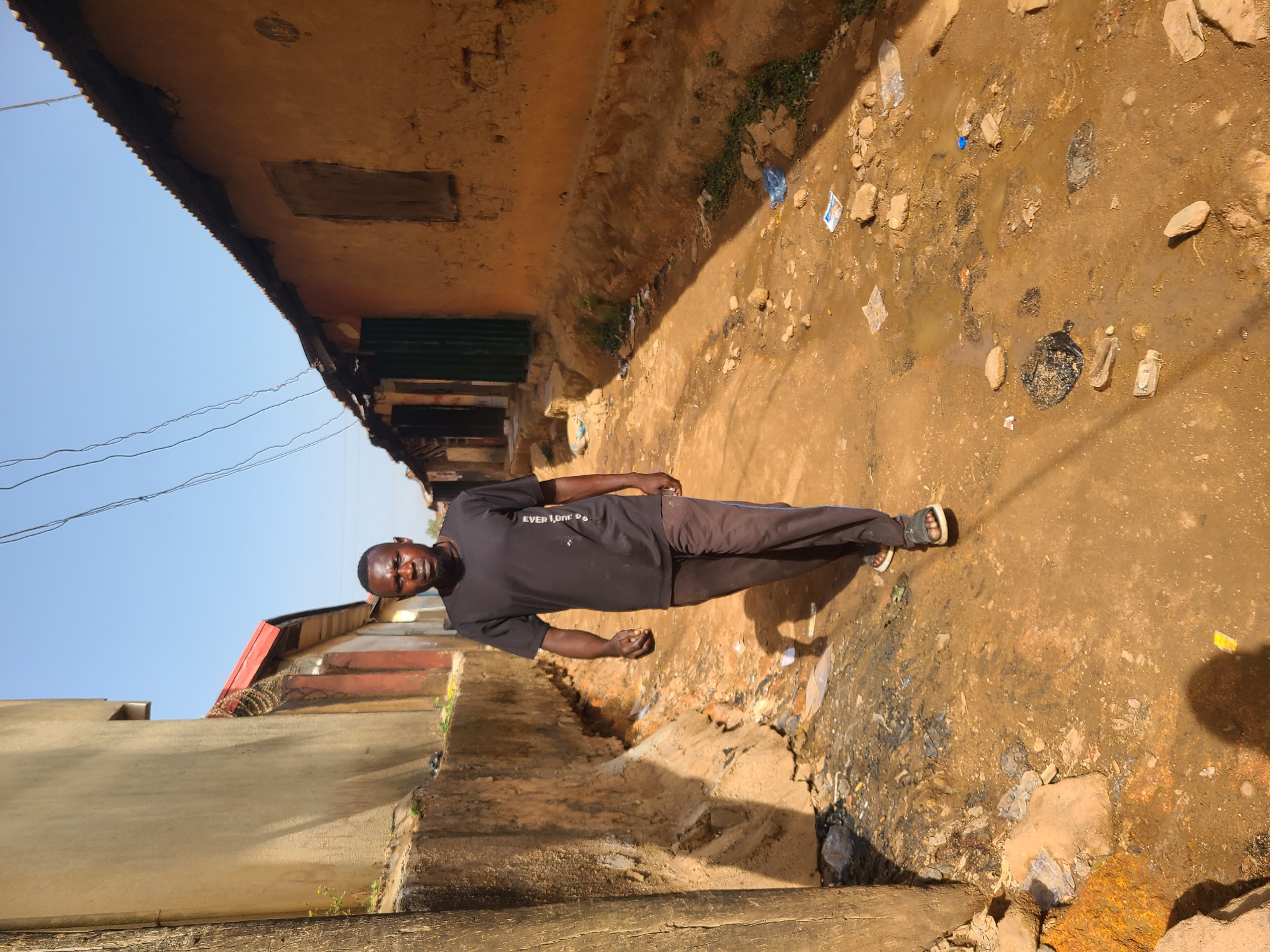 An outdoor, eye-level shot depicts an adult male standing in a narrow, unpaved alleyway or pathway in what appears to be a residential area. The image has been rotated and shows the man standing upright relative to the ground.
The man, of African descent, is centrally positioned, looking directly at the camera with a neutral to slight smiling expression. He wears a black t-shirt with partially visible white text that reads "EVER LOR" (likely "EVER LORD'S"), dark trousers, and open-toed sandals. His short hair and beard are dark.
The ground around him is uneven, muddy, and littered with various debris, including plastic bottles, wrappers, stones, and what appears to be stagnant water or small puddles. This suggests poor drainage and sanitation conditions.
To the viewer's left, a light-colored concrete wall forms the side of a building or boundary. To the right, an older, simpler building with a corrugated metal roof and a faded, possibly mud-colored wall is visible. Dark green structures, possibly doors or shutters, are present on this building. Further in the background on the right, another building with a reddish-brown roof can be seen.
The sky above is clear and bright blue, indicating sunny weather during the day, likely midday or afternoon due to the strong light. Thin electrical wires crisscross the sky. The overall scene suggests a humble, possibly impoverished, urban or semi-urban setting, consistent with its context in Jos, Nigeria.