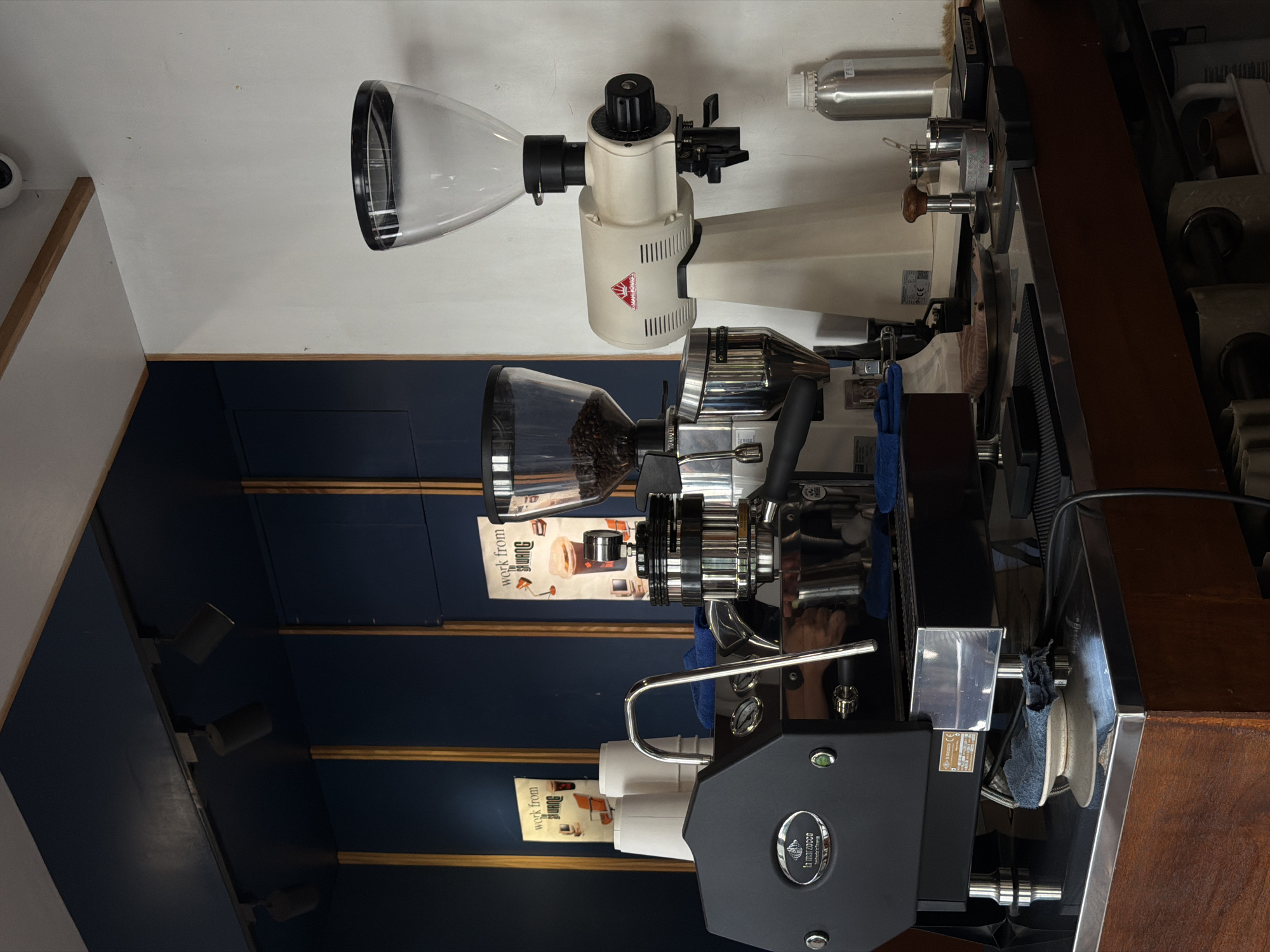 This is a medium shot, taken from a low angle, of a coffee shop counter in Chiang Mai City Municipality, Thailand. The counter is made of dark wood and stainless steel. On the counter are a black espresso machine with a La Marzocco logo, a white Mahlkönig coffee grinder, and two posters on the dark blue wall behind the counter. The posters advertise "work from Buang" and feature images of coffee cups. The overall lighting is dim, with spotlights focused on the counter area. There are no people visible in the image.