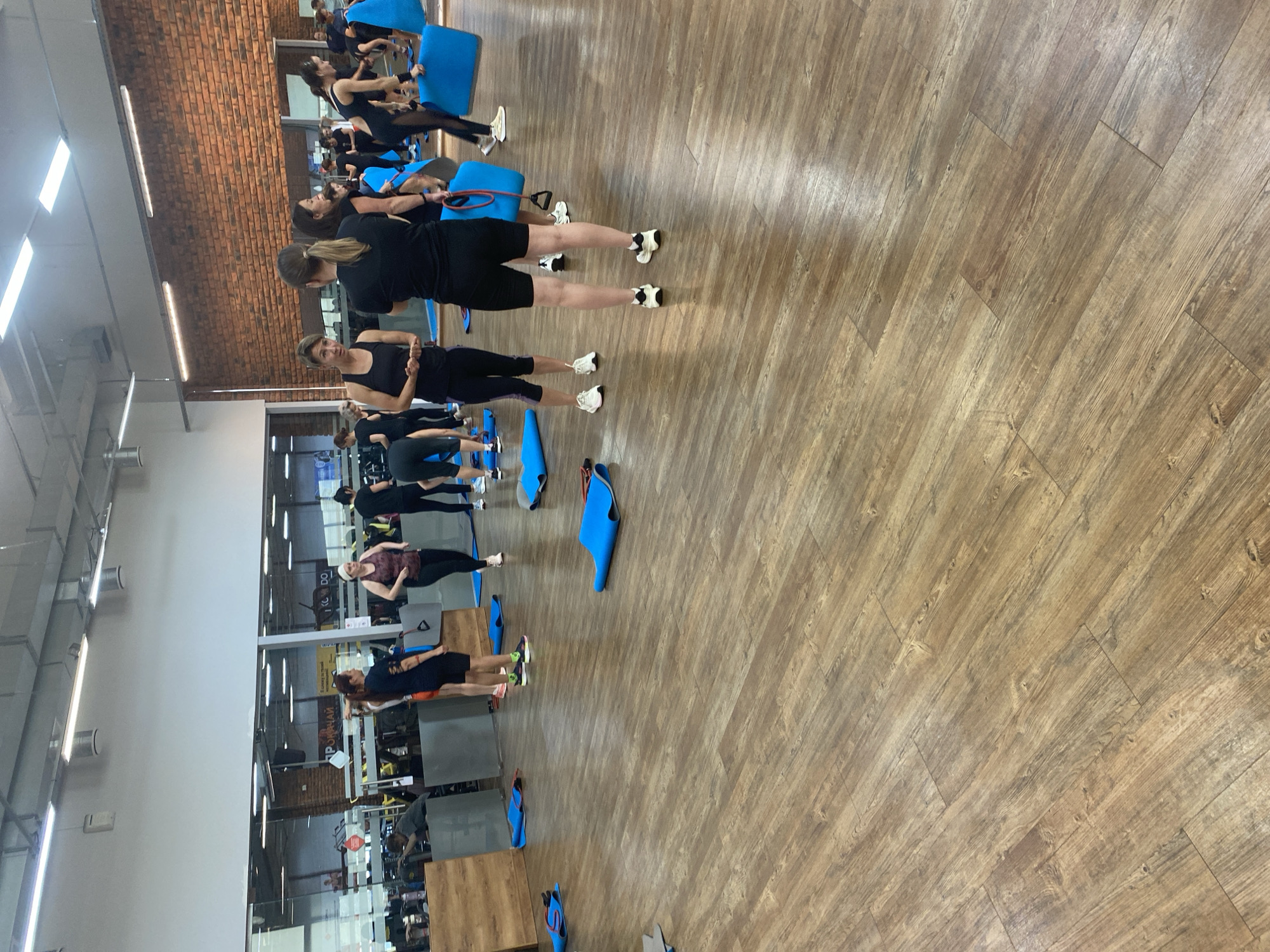 The image depicts an indoor fitness class taking place in a gym. Several individuals, mostly women, are dressed in athletic attire and appear to be participating in or preparing for an exercise routine. Blue yoga mats are spread across the wooden floor. In the background, gym equipment and mirrors are visible, and one wall is covered in exposed brick. The lighting appears to be from overhead fluorescent fixtures.