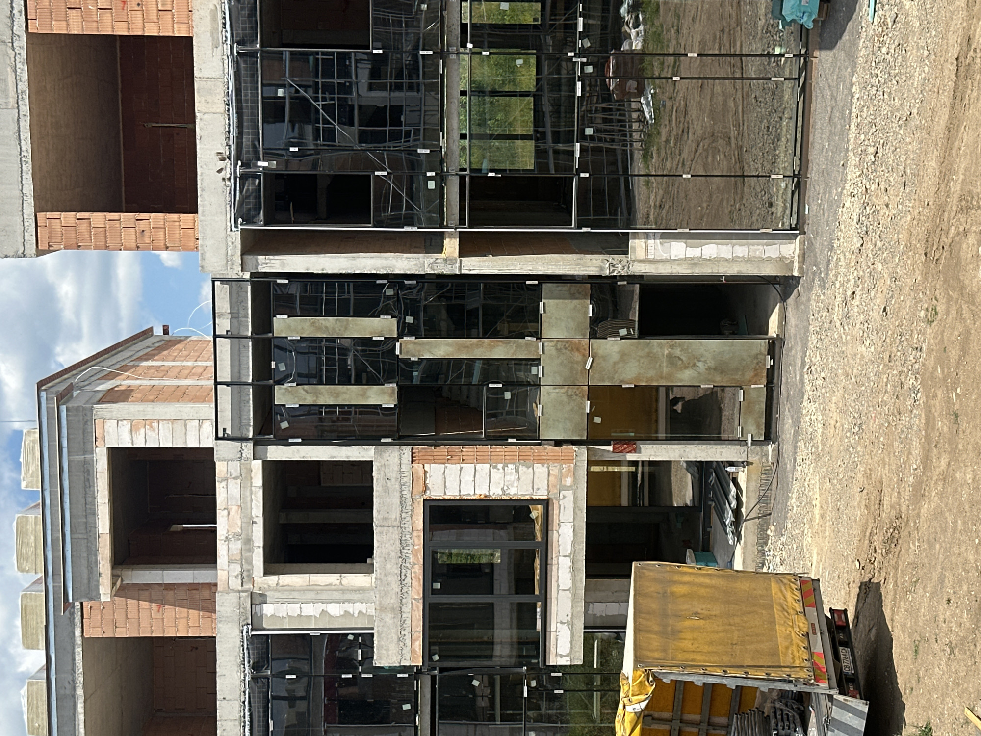 This is a vertical, outdoor, medium shot of a multi-story building under construction in Stara Zagora, Bulgaria. The building's facade is a mix of exposed brick, concrete, and large, dark-tinted glass windows framed by black metal. Some windows are partially covered with protective white stickers. The building appears to be several stories high, with open, unfinished interior spaces visible through some of the window openings.
To the right of the building, a dirt and gravel area is visible, suggesting a construction site. A yellow-covered truck is parked on the gravel, its rear end facing the camera. The truck has a license plate that reads "CB 7714 CA".
The sky is blue with scattered clouds, indicating fair weather. The time of day appears to be daytime, likely morning or afternoon, given the lighting and shadows. The overall scene depicts an active construction project.