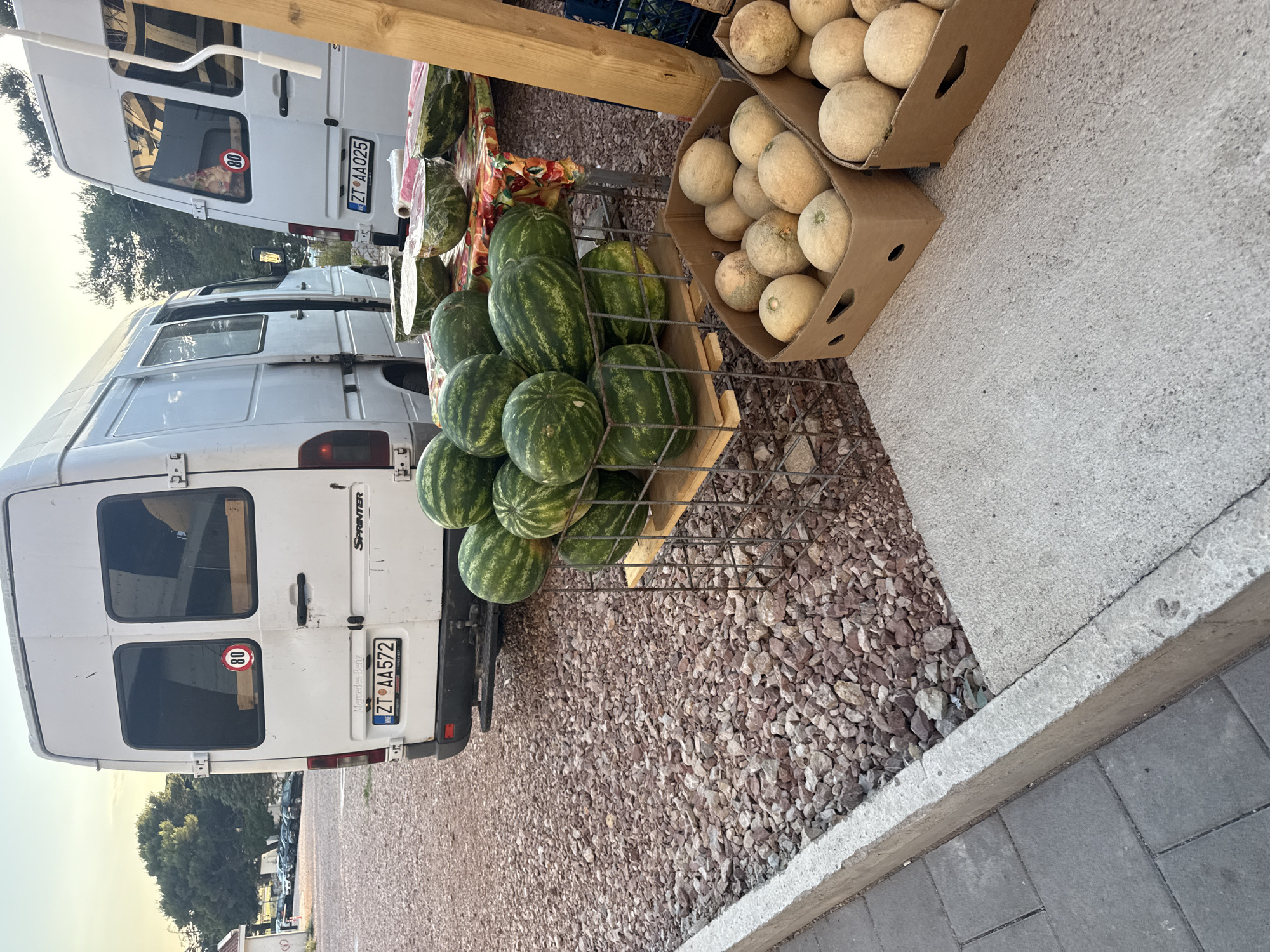 The image depicts an outdoor market scene in Rafailovići, Montenegro, with two white Mercedes-Benz Sprinter vans parked side-by-side. The van on the left has a license plate reading "ZT AA572". The van on the right has a license plate reading "ZT AA025". In front of the vans, a variety of fruits are displayed for sale. A metal rack holds a collection of whole watermelons, some of which are partially wrapped in plastic. To the right of the watermelons, two cardboard boxes are filled with cantaloupes. A table draped with a colorful tablecloth is also visible, displaying more cut and whole watermelons, some of which are also wrapped in plastic. The ground is covered with gravel and a concrete walkway borders the market area. The sky suggests it is either early morning or late afternoon. There are no people visible in the image.