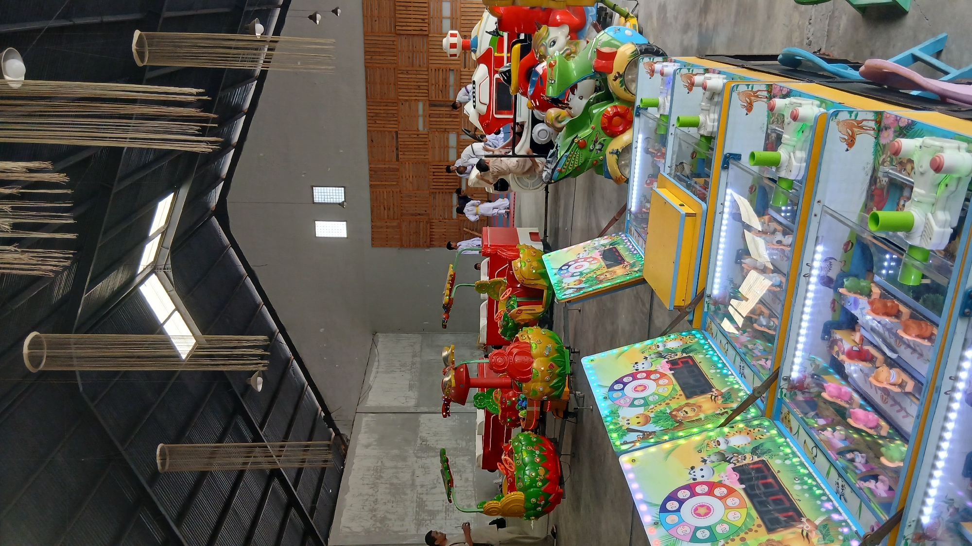 The image depicts an indoor children's play area, likely an arcade or amusement center, located in the City of Medan, Indonesia. The space features a variety of colorful, coin-operated rides for children, including carousel-style units shaped like fruit and cartoon animals, and a larger red and white vehicle-shaped ride. Several children, some wearing white martial arts uniforms with yellow belts, are visible interacting with the rides and a central mat area. The environment is a large, open hall with a high, corrugated metal roof and visible skylights. Decorative, rope-like hanging installations are suspended from the ceiling. In the foreground, several electronic game machines with bright displays and small prize compartments are arranged on a yellow and blue counter. The overall impression is a lively and engaging space designed for children's entertainment.