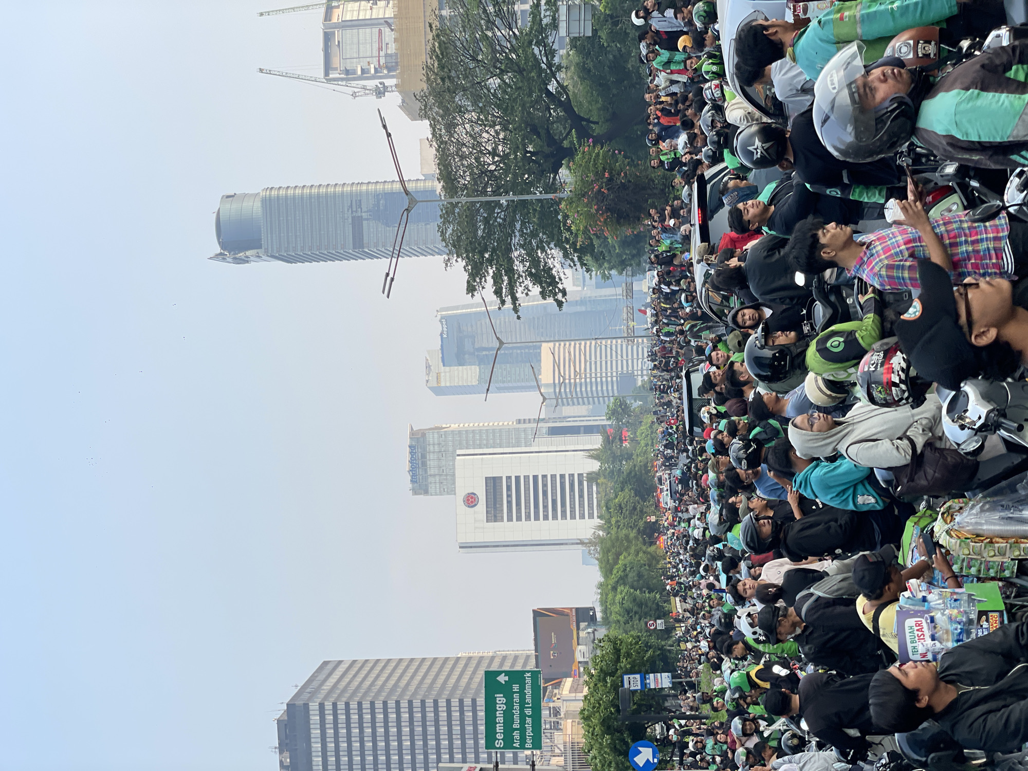 A wide shot captures a street scene in Central Jakarta, Indonesia, characterized by a significant traffic jam and a large gathering of people, many of whom are on motorcycles. The sky is hazy, suggesting it might be dawn or dusk, or perhaps air pollution. In the background, several modern skyscrapers dominate the skyline, including a distinctive tall, curved glass building. Construction cranes are visible, indicating ongoing development.
A prominent green traffic sign on the left reads "Semanggi Arah Bundaran HI Berputar di Landmark," indicating directions to Semanggi, HI Roundabout, and a landmark. Below this sign, a blue directional arrow points left. Other traffic signs, including a "STOP" sign, are also present.
The majority of the people in the foreground and middle ground are wearing casual clothing and many have helmets on, indicative of motorcycle riders. A significant number are wearing green jackets or vests, possibly indicating they are delivery riders or part of a ride-sharing service. They are gathered on and around their motorcycles, creating a dense crowd on the road. Some individuals are seen interacting with each other, while others appear to be waiting. Several motorcycles are lined up, contributing to the traffic congestion. In the lower foreground, a vendor is visible with a display of bottled drinks and snacks. The overall impression is of a busy urban environment during a traffic standstill, with a large concentration of motorcyclists.