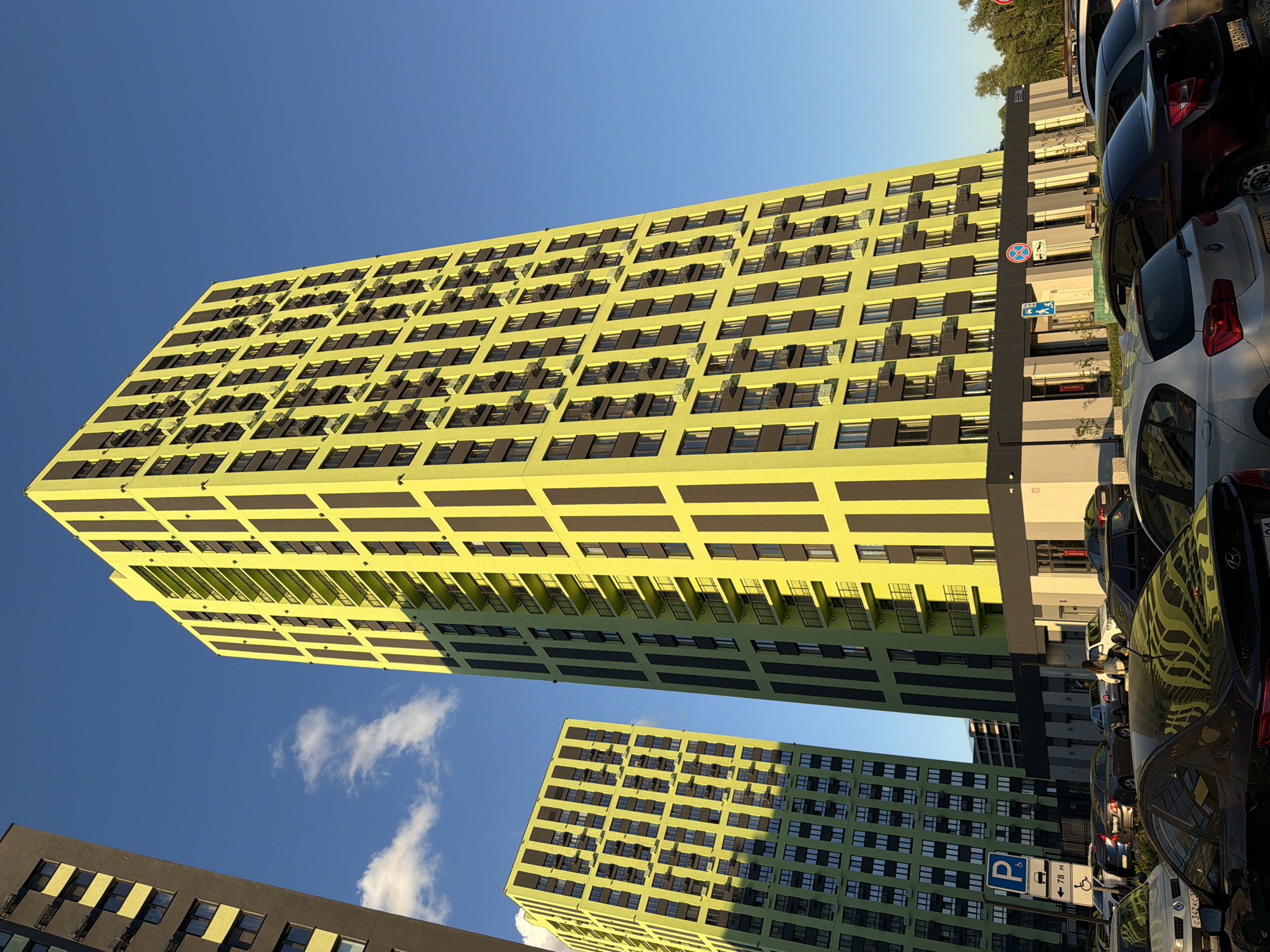 In the city of Unknown, Unknown, two contemporary high-rise apartment buildings dominate the frame under a clear blue sky. The building on the right is taller and has a striking lime green facade with dark vertical stripes, numerous balconies, and rows of windows. To its left, a shorter, similarly designed building also features a lime green facade with dark accents. A dark-colored building is partially visible on the far left. In the foreground, a parking lot is filled with various cars, with some parked in front of the buildings. Several street signs are visible near the entrance of the buildings, including a blue parking sign with an accessibility symbol and a red circular sign with a diagonal bar. The sun casts long shadows across the scene, indicating it is either early morning or late afternoon. There are a few wispy clouds in the sky.