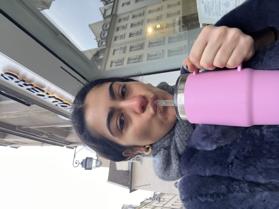 An individual, identified as an adult female, is depicted in an outdoor close-up on a street in Paris, France. The visible "VOLKERS" sign above her, consistent with VOLKERS & NANEUIL located at 1 Rue du Pont Louis Philippe, 75004 Paris, identifies the specific urban location. She wears a dark blue, possibly faux fur, coat over a grey scarf, and has golden hoop earrings. Her dark hair is pulled back. She holds a large, pink travel mug with a handle and a metallic lid in her right hand, actively drinking from it through a clear straw, her lips visibly pursed. Her fingernails are painted dark blue. The background features a building facade with the illuminated "VOLKERS" sign, a large storefront window reflecting a multi-story Haussmannian-style Parisian building, and an older stone building to the left, adorned with a traditional black ornate street lamp. The sky is overcast.