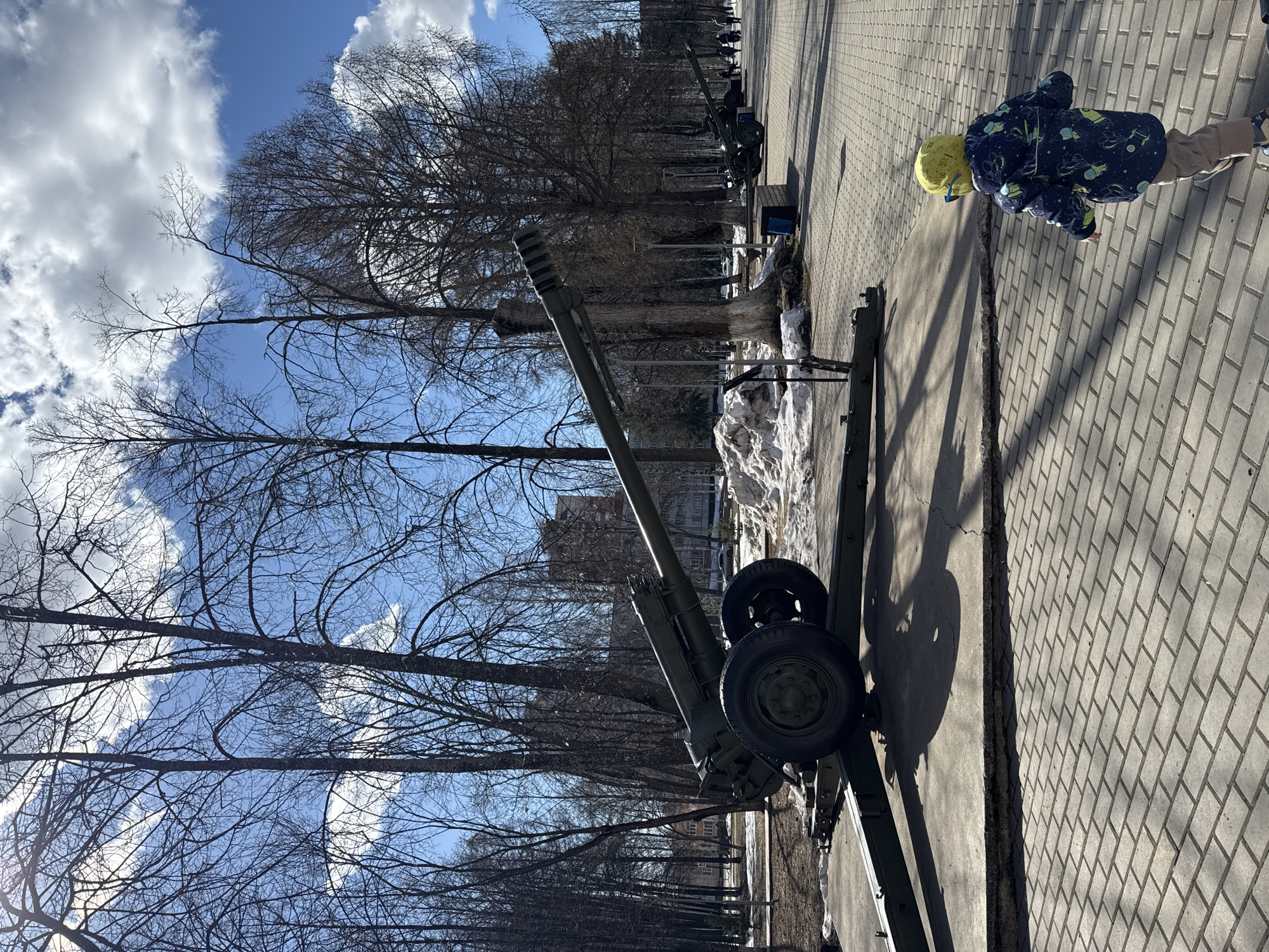 A child in a blue and green patterned jacket and yellow hat is looking at the ground on a paved surface. An artillery gun is in the foreground on the left, pointing towards the sky. Trees are visible behind the gun, and patches of snow are on the ground nearby. Other guns are lined up in the distance along the paved surface. The sky is blue with scattered clouds. The location is Izhevsk, Russia, as evidenced by the artillery guns which are likely a part of a military memorial or display.