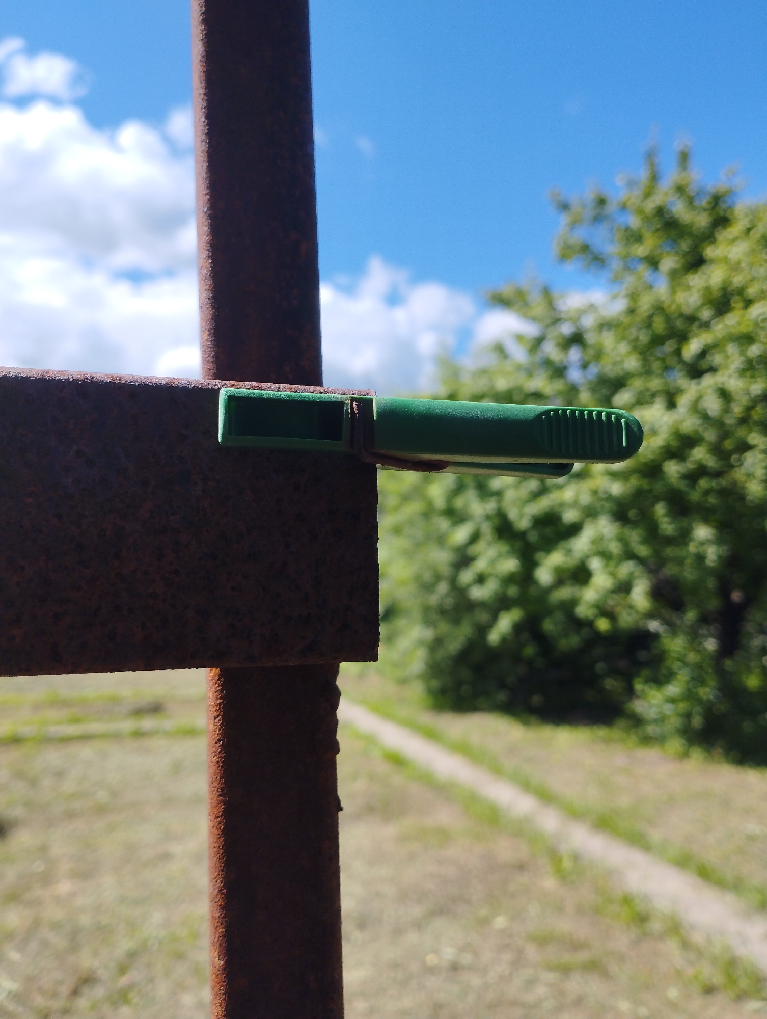 The image shows a close-up of a rusty metal pole with a green clothespin attached to a horizontal metal bar. In the background, there is a blurred view of green trees, a dirt path, and a blue sky with white clouds. The scene appears to be outdoors, likely in a rural or park-like setting. The specific location of Ширяево (ильинский), Russia, is not visually discernible from the image itself. There are no people, actions, or interactions depicted.