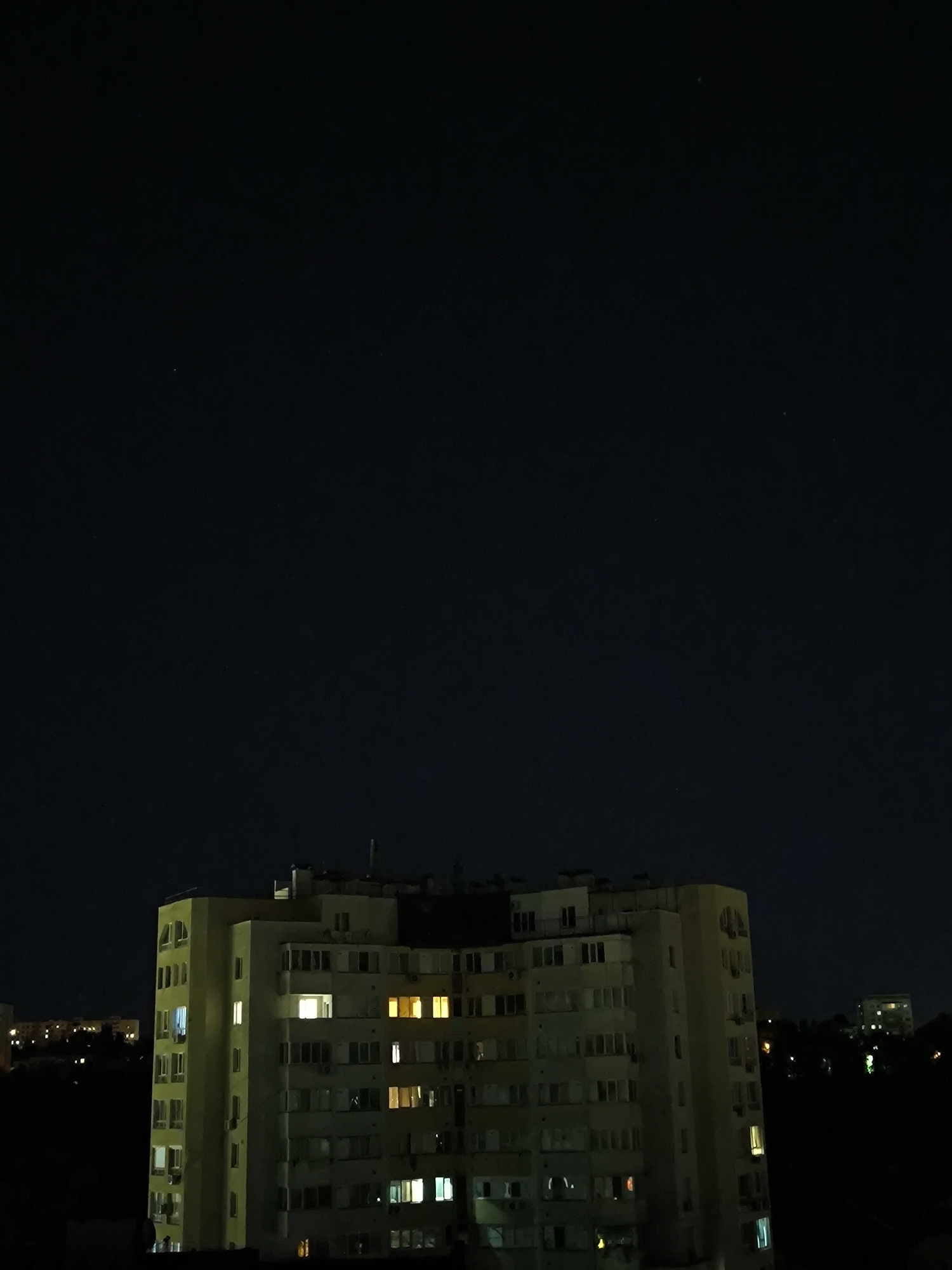 A night-time photograph captures a tall, multi-story apartment building in Chișinău, Moldova. The building, primarily yellow and off-white, features numerous windows, many of which are illuminated with warm interior lights, suggesting occupancy. The architectural style appears consistent with Soviet-era housing blocks, common in Eastern European cities. The building is situated within an urban landscape, with other, less distinct buildings visible in the background, also showing signs of illumination. Trees and dark, shadowed areas suggest a natural environment surrounding the built structures. The sky above is a deep, dark blue, indicative of a clear night, with a few faint stars visible. The overall impression is one of a quiet urban scene at night.