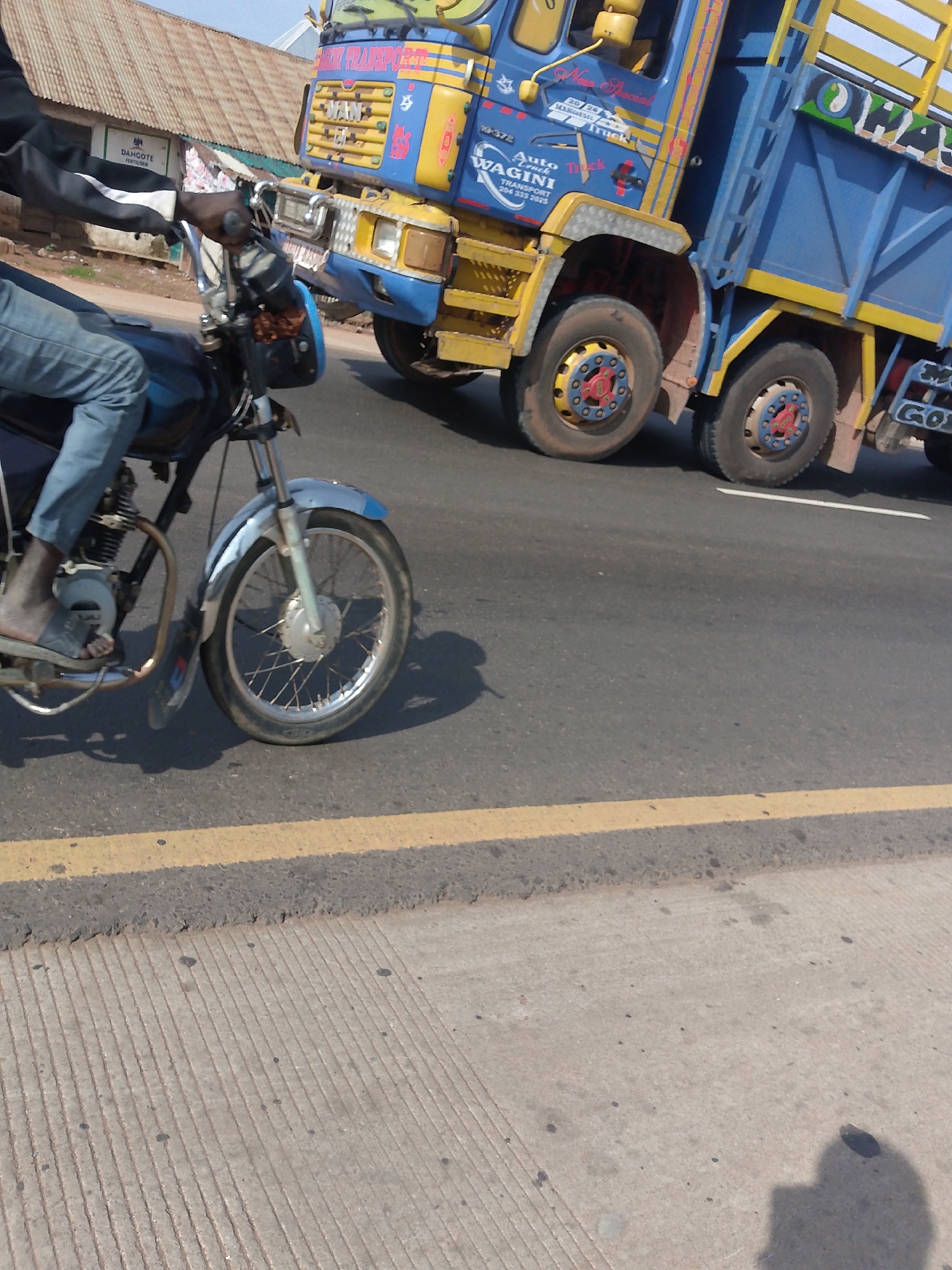 This is a street-level, eye-level, medium shot of a motorcyclist in the foreground and a large blue and yellow truck in the background. The scene appears to be in Keffi, Nigeria. The motorcyclist, wearing blue jeans and a dark jacket, is riding a motorcycle with a silver front fender and spokes. The motorcycle's engine is visible. The truck is a multi-wheeled vehicle with a yellow cab and a blue cargo bed, adorned with colorful graphics and text, including "MAN" and "WAGINI TRANSPORT." The truck has double wheels on its rear axles. The road surface is asphalt with a yellow line dividing lanes. A corrugated metal roof structure and a sign that reads "DAHCOTE" are visible on the left side of the image, suggesting a commercial or residential area. The lighting indicates it is daytime.