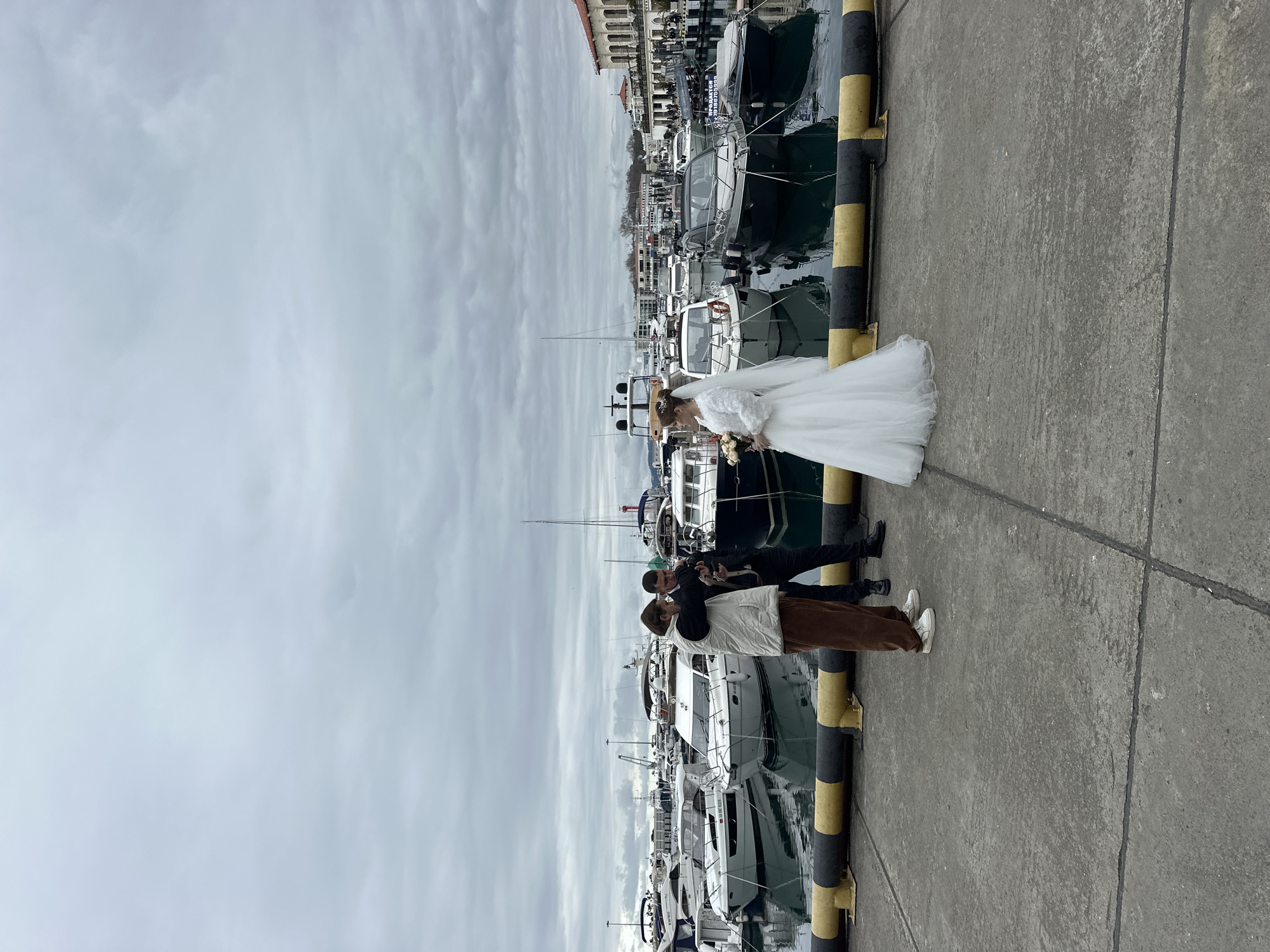 The image displays an outdoor scene at the Sochi Sea Port (Sochi Grand Marina) in Sochi, Russia, under an overcast sky. In the foreground, a concrete pier extends across the frame, with yellow and black striped safety bollards marking its edge along the water. Three individuals are positioned on the pier. A woman, dressed in a white wedding gown with a veil and holding a bouquet of light-colored flowers, stands facing left, looking towards the water. To her left, two men are standing close together, looking down at a device held by one of them. The man holding the device is wearing a dark suit, while the other man is dressed in a white sleeveless vest over a lighter top, brown pants, and white sneakers. The background is dominated by a marina filled with numerous docked motor yachts and smaller boats, some featuring visible Russian markings like "ЯХТ-КЛУБ 'АДЛЕР'". Beyond the marina, a large, multi-story building with classical architectural features, including arches and red-tiled roofs, consistent with the Sochi Sea Port terminal, is visible. The water in the marina is dark and reflective.