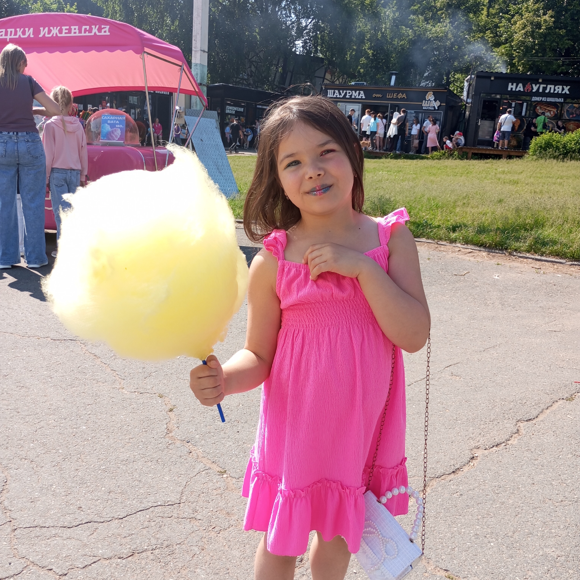 In Izhevsk, Russia, a young girl stands outdoors holding a large ball of yellow cotton candy. She is wearing a bright pink dress with ruffled straps and hem, and a small white purse with a pearl strap is hanging from her shoulder. Her lips are adorned with blue and silver glitter. Behind her, a pink tent displays the text "СЛАДКИЕ ИЖЕВСКА" (Sweet Izhevsk), and a sign inside reads "САХАРНАЯ ВАТА" (Cotton Candy). Several food trucks, including one with "ШАУРМА ОТ ШЕФА" (Shawarma from the Chef) and another labeled "НА УГЛЯХ" (Over Coals) selling Doner Kebab, are visible in the background. People are seen queuing at the food trucks. The ground is paved asphalt with visible cracks, and a grassy area with trees forms the backdrop under a bright, sunny sky.