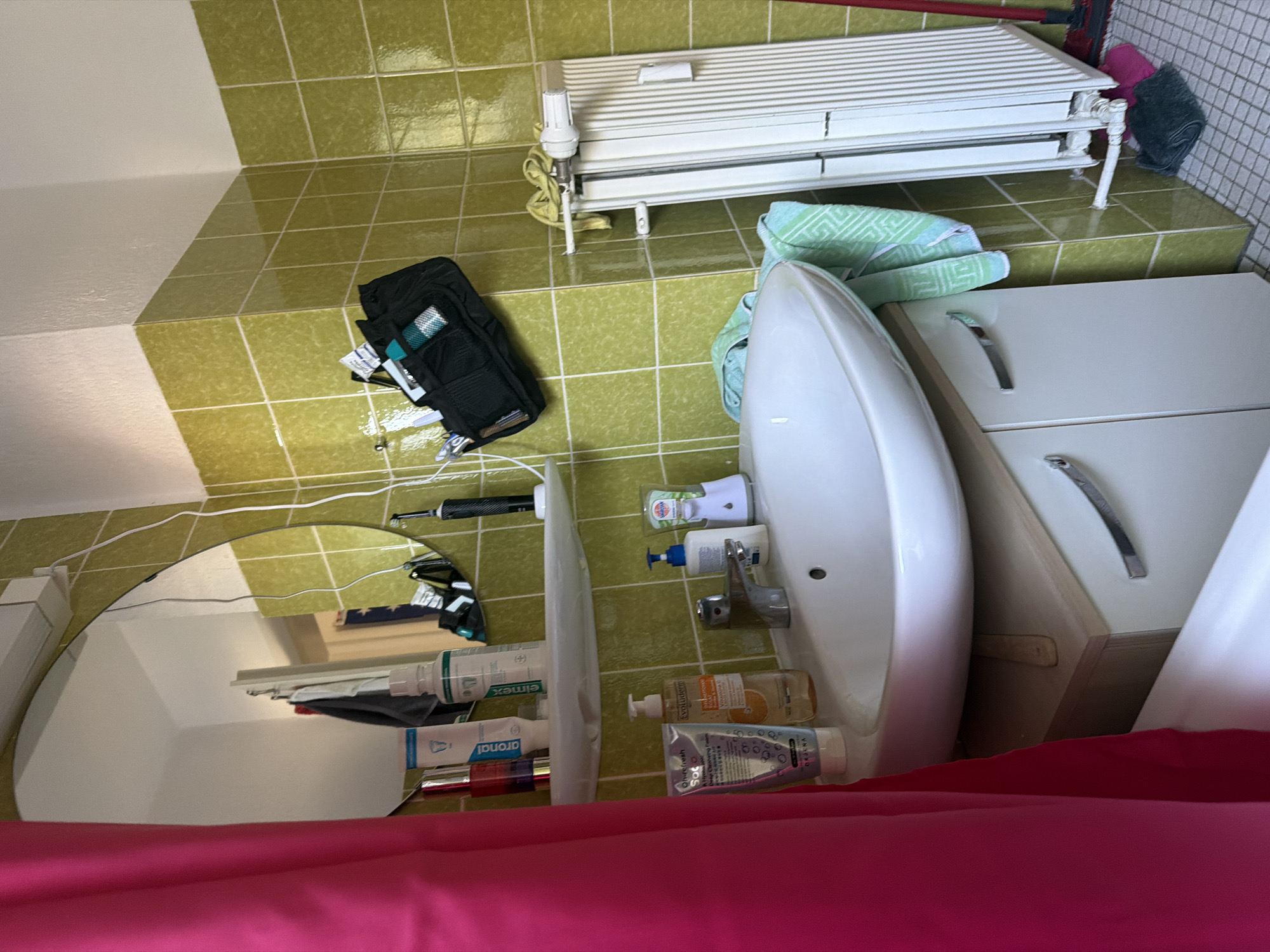 The image depicts a bathroom interior, likely in a residential setting. The walls are tiled with a lime-green, glossy finish, creating a vibrant and somewhat dated aesthetic. On the left, a bright pink shower curtain is visible, partially obscuring the view. A white sink with a chrome faucet is the central focus of the lower part of the image. Above the sink, a white shelf holds various toiletries, including bottles of mouthwash and toothpaste, and a red bottle, possibly perfume. A black toiletry bag is hung on the wall above the shelf, containing several items. To the right of the sink, a white radiator is mounted on the wall. The floor appears to be tiled with small, light-colored squares. On the floor near the radiator, a dark-colored cloth and a pink item are present, along with a red mop. The overall impression is a functional, albeit slightly cluttered, bathroom environment.
Given that the request specifies Nienburg/Weser, Germany, this scene could be a bathroom within a typical apartment or house in that city. Such bathrooms are common in older residential buildings in Germany, characterized by tiled walls and radiators for heating. The specific placement of the items suggests everyday use, with toiletries readily accessible near the sink. The presence of a radiator is a standard heating fixture in German homes.