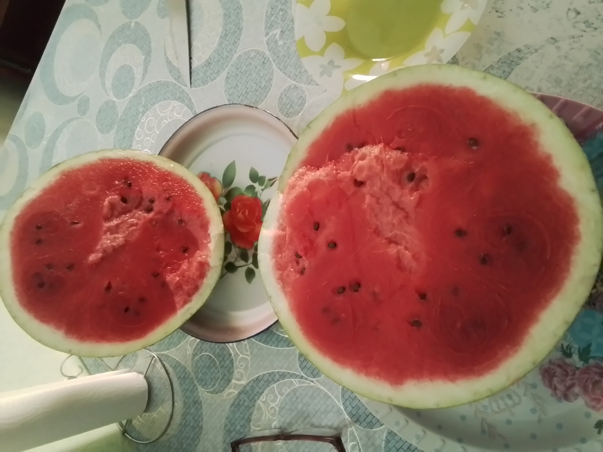 A top-down, medium shot captures two halves of a watermelon resting on a patterned tablecloth. The watermelon is bright red with visible black seeds and a light green rind. One half of the watermelon is positioned on a white plate with a floral design, while the other half is placed directly on the table to the right of it. In the background, a roll of paper towels stands on a metal holder. The scene is set indoors, likely in a kitchen or dining area, with the patterned tablecloth suggesting a domestic environment. No specific location in Taraz, Kazakhstan, can be identified from the image alone.