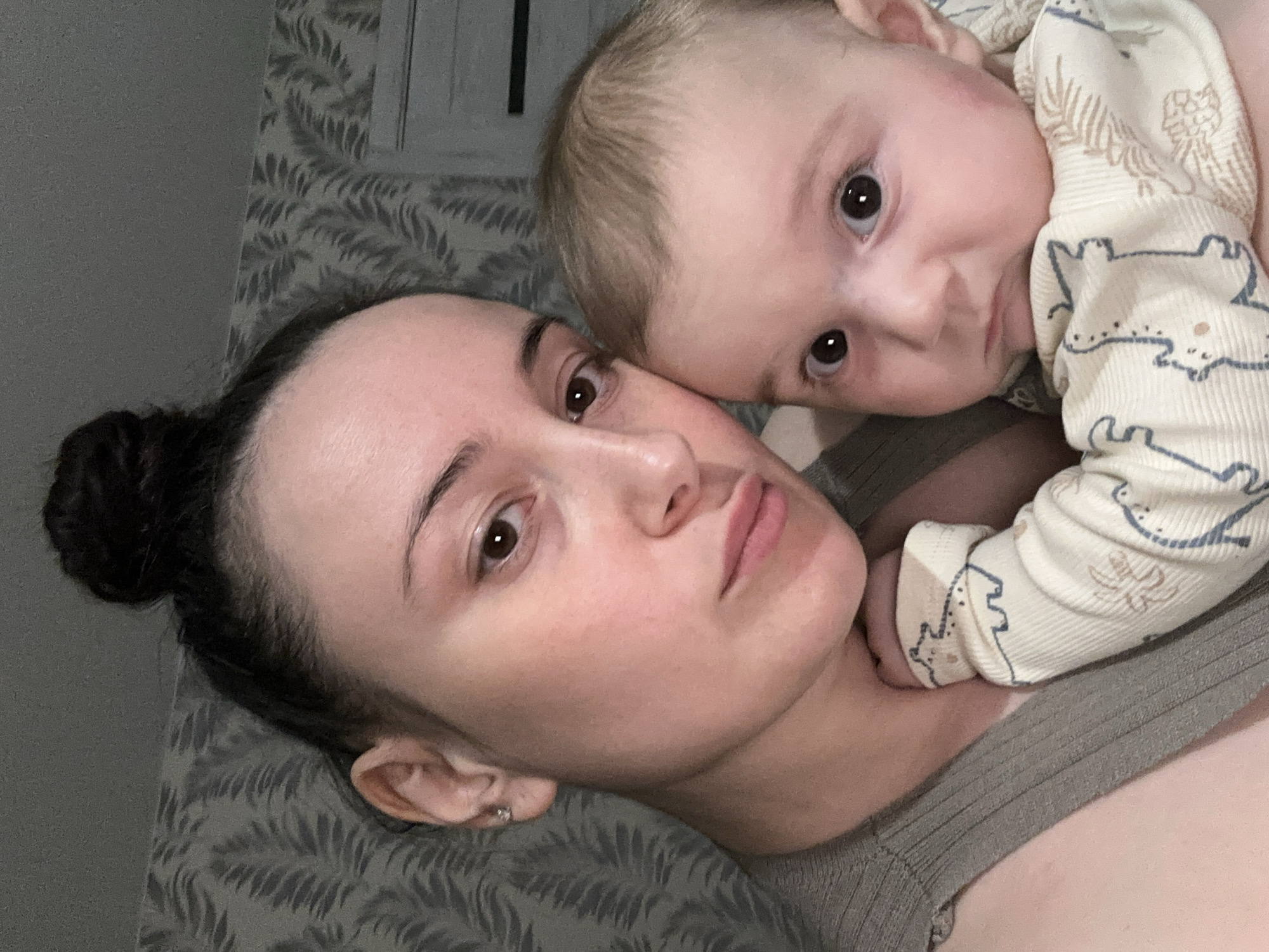 A woman with her hair in a bun lies on a bed or surface covered in grey patterned fabric, a young child lies on top of her. The woman looks directly at the camera with a neutral expression. The child, in a patterned white onesie, also looks at the camera. The environment is indistinct but appears to be indoors. It is not possible to confirm the location is Chaikovsky, Russia.