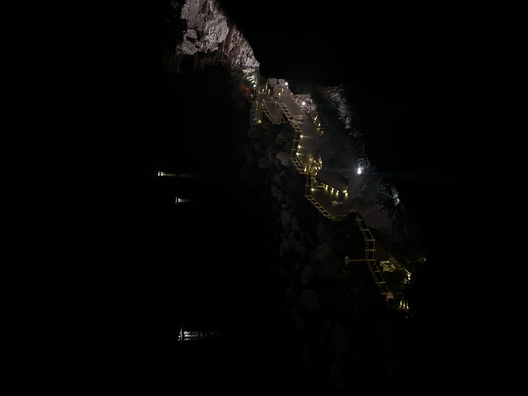 A nighttime, aerial view captures a well-lit wooden boardwalk winding along a rocky coastline. The boardwalk is illuminated by numerous small yellow lights, creating a pathway that curves and ascends the rocky terrain. Several individuals are visible walking on the boardwalk. In the distance, on the dark water, there are faint lights from what appear to be boats. The rocky shoreline is rugged and dark, with some areas of the cliff face illuminated by the boardwalk lights. The scene suggests an outdoor recreational area or scenic overlook. This location is the Singing Dunes (also known as Bozzhyra tract) near Aktau, Kazakhstan.