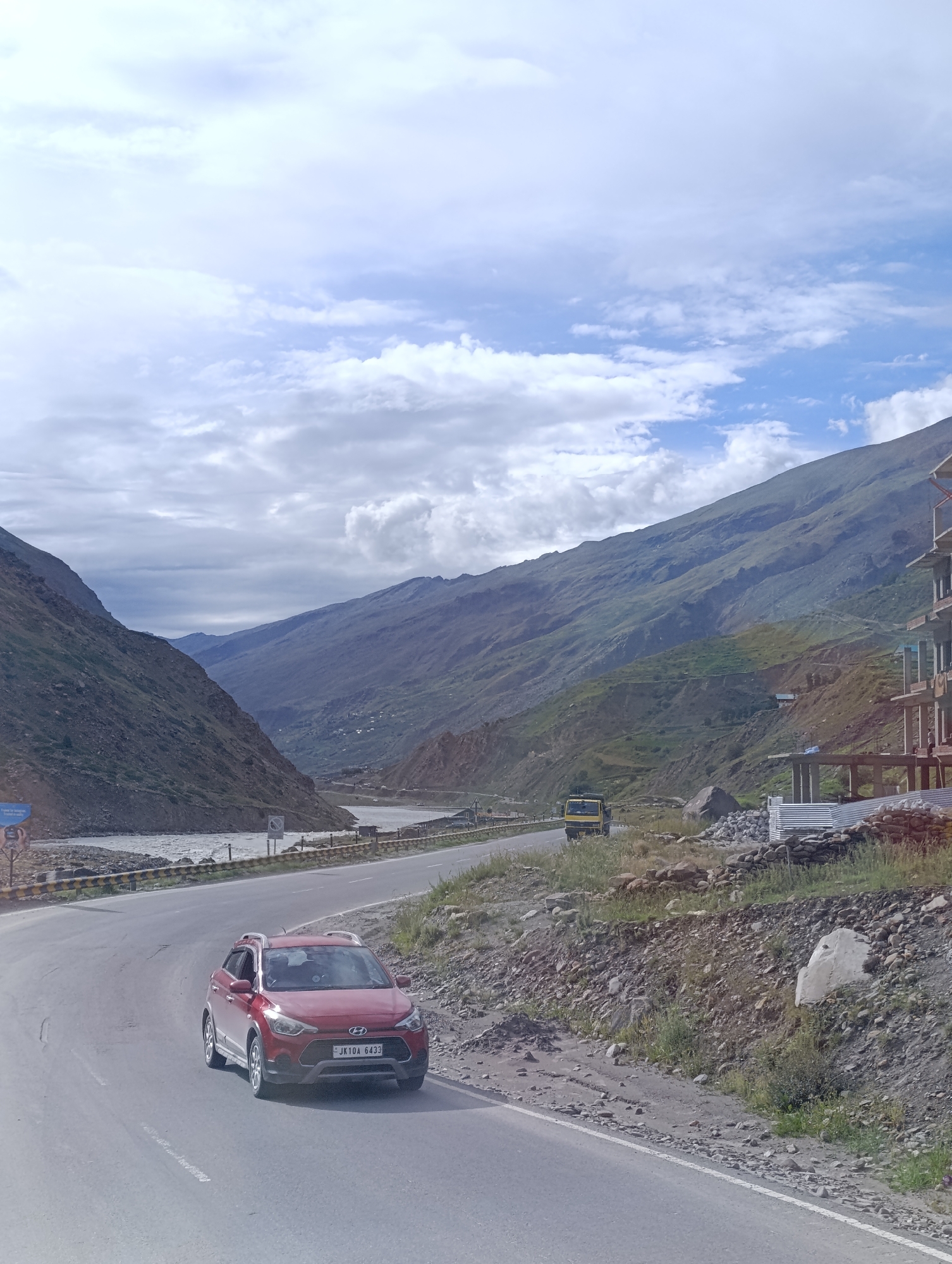 The image depicts a winding road in a mountainous region, likely near Sissu, India, as suggested by the location context. A red Hyundai i20 car, with license plate "JK10A 6433", is prominently featured on the right side of the road, traveling away from the viewer. The car appears to be in motion.
The road curves to the left, with a steep, rocky mountain slope on the right. The left side of the road overlooks a river that flows through a valley. Further in the distance, more mountains rise, displaying layers of green and brown hues, with some signs of human settlement visible on the slopes. The sky is partly cloudy, with a mix of white and grey clouds against a pale blue background.
On the right side of the road, there are some construction materials, including corrugated metal sheets and piles of stones, suggesting ongoing development or infrastructure work. A yellow truck is visible in the distance, also on the road, heading in the same general direction as the red car. The overall environment is natural and rugged, characteristic of a Himalayan landscape. There are no visible people interacting with each other or the environment in a significant way.