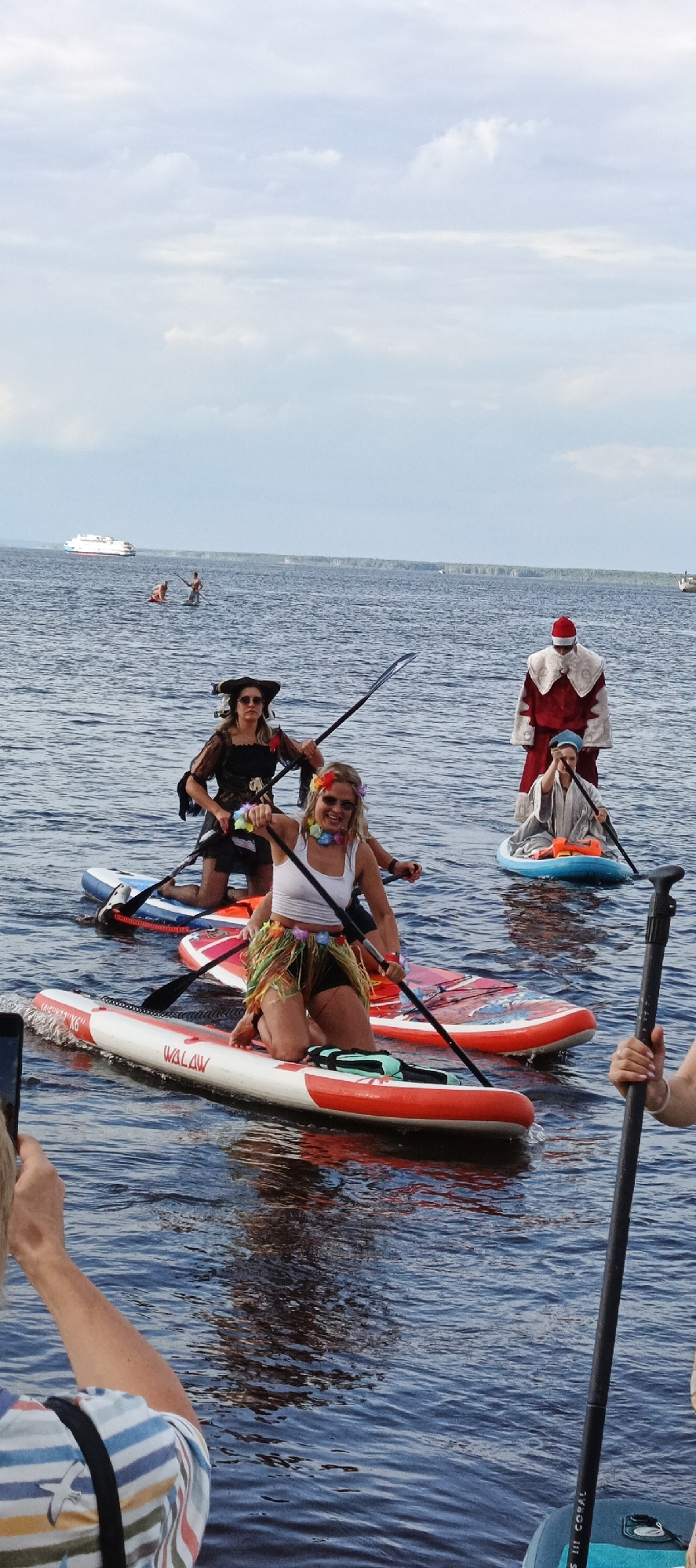 The image depicts an outdoor event on a body of water, possibly a river or lake, under a cloudy sky. In the foreground, several people are paddleboarding. One person in the center is dressed in a white top and a grass skirt, kneeling on a red and white paddleboard. To their left and slightly behind, another person is dressed as a pirate, also kneeling on a paddleboard. Further back and to the right, someone is dressed in a Santa Claus costume with a red and white coat and hat, standing on a blue paddleboard. Two other individuals are visible in the background, one appearing to be in the water near a paddleboard, and another standing on a paddleboard. A large white ferry or ship is visible in the distance on the water, with a treeline on the horizon. The scene suggests a recreational event or festival taking place on the water. The location is identified as Chaikovsky, Russia.