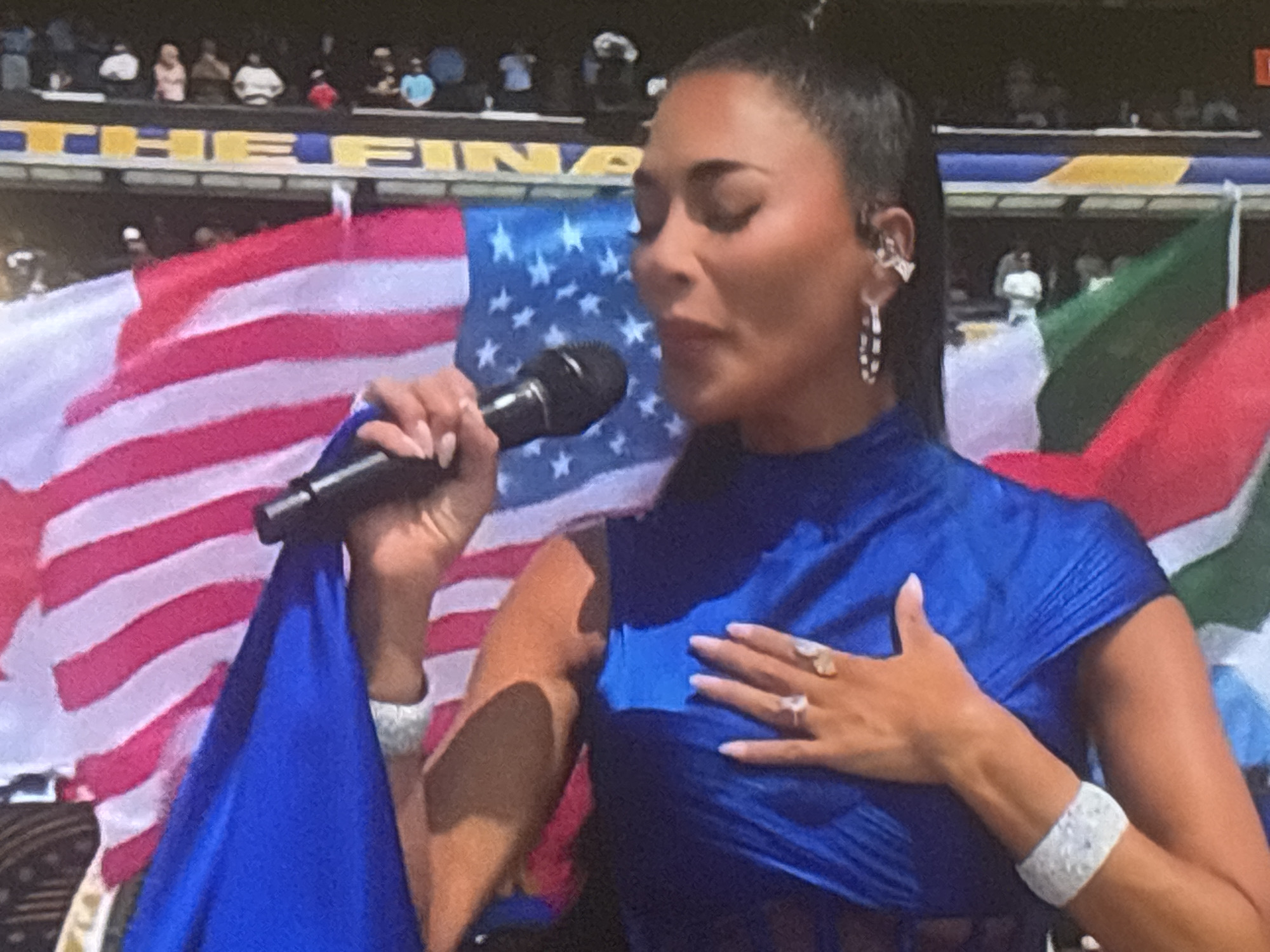 A woman with dark hair pulled back is singing into a microphone, her right hand is on her chest, wearing a blue top and a white diamond bracelet. She has rings on her fingers, and she is wearing a large earring on her left ear. Behind her, an American flag is visible, as well as other flags. In the background, a crowd is visible, and a stadium banner displays the word "FINALS". The scene is set in a stadium.