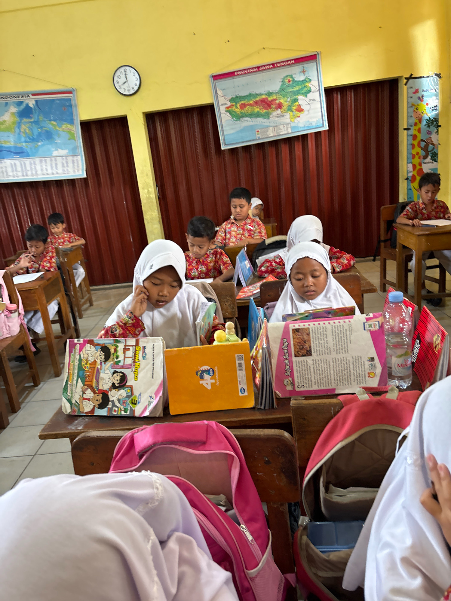The image depicts a classroom scene at a school in Tirtomoyo, Indonesia. Several young students, mostly appearing to be of elementary school age, are seated at wooden desks, engaged in learning activities. The room is brightly lit with yellow walls and has a somewhat rustic appearance, with maroon-colored corrugated metal doors visible in the background.
In the foreground, a girl wearing a white hijab and a white uniform with red and white patterned sleeves is focused on a book in front of her. She is holding her hand to her ear as she reads. Beside her are several books and an orange folder. To her right, a pink backpack is open, revealing contents. Further to the right, another student is partially visible, also wearing a white hijab.
Other students are visible in the mid-ground and background. Some are wearing similar white uniforms, while others are in red and white patterned shirts. They are also engrossed in their work, with books and papers on their desks. A clock hangs on the wall above the students, showing approximately 9:25. Maps and educational posters are displayed on the walls, including a map of Indonesia and a regional map labeled "PROVINSI JAWA TENGAH". A height chart with cartoon animals is also visible on the right wall. The overall impression is one of a typical school day in a primary education setting.